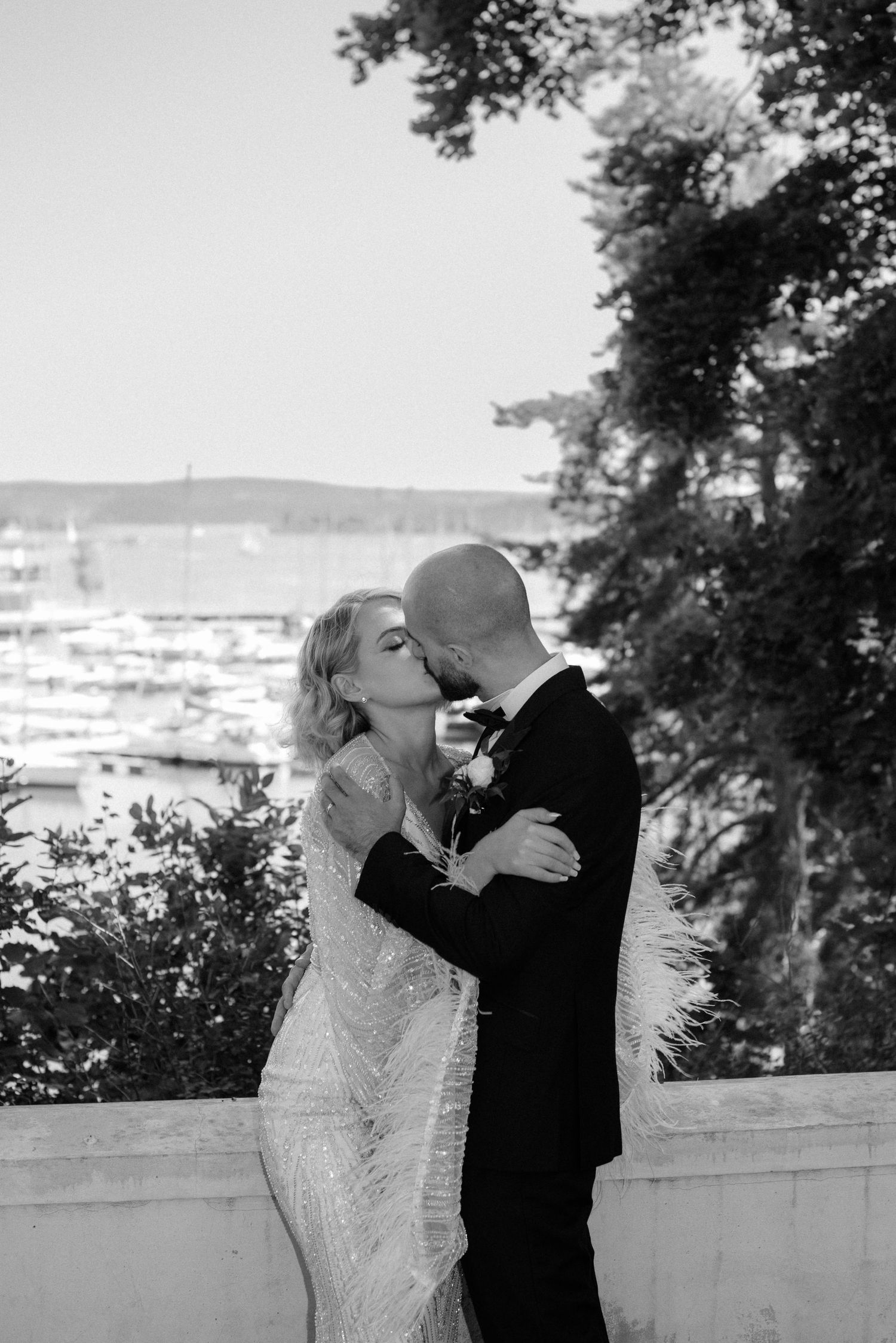 A romantic couple shares a kiss at an overlook with a marina and boats visible in the background.