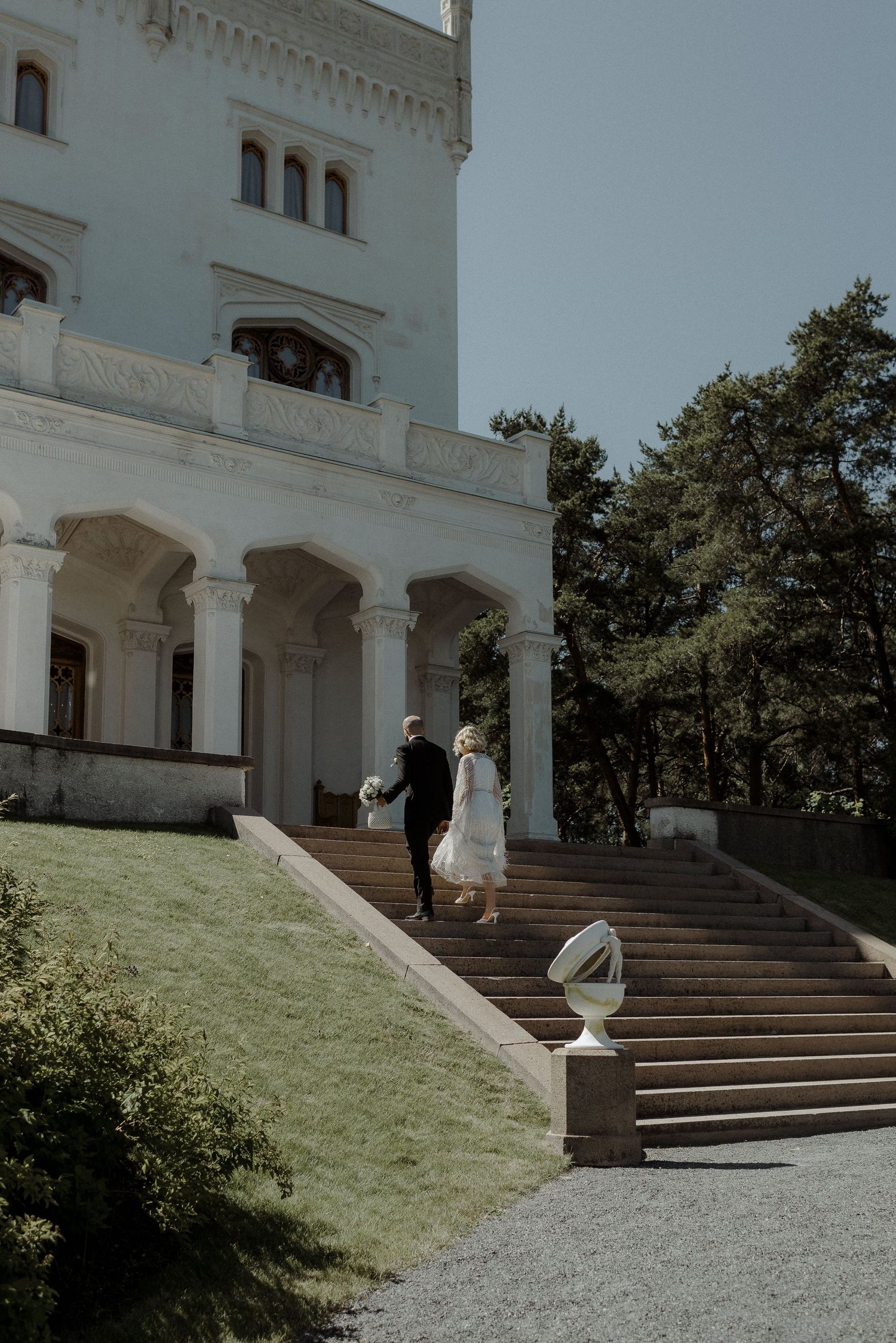 A couple stands on the grand stone staircase of a white mansion with ornate architecture and lush grounds on a sunny day.