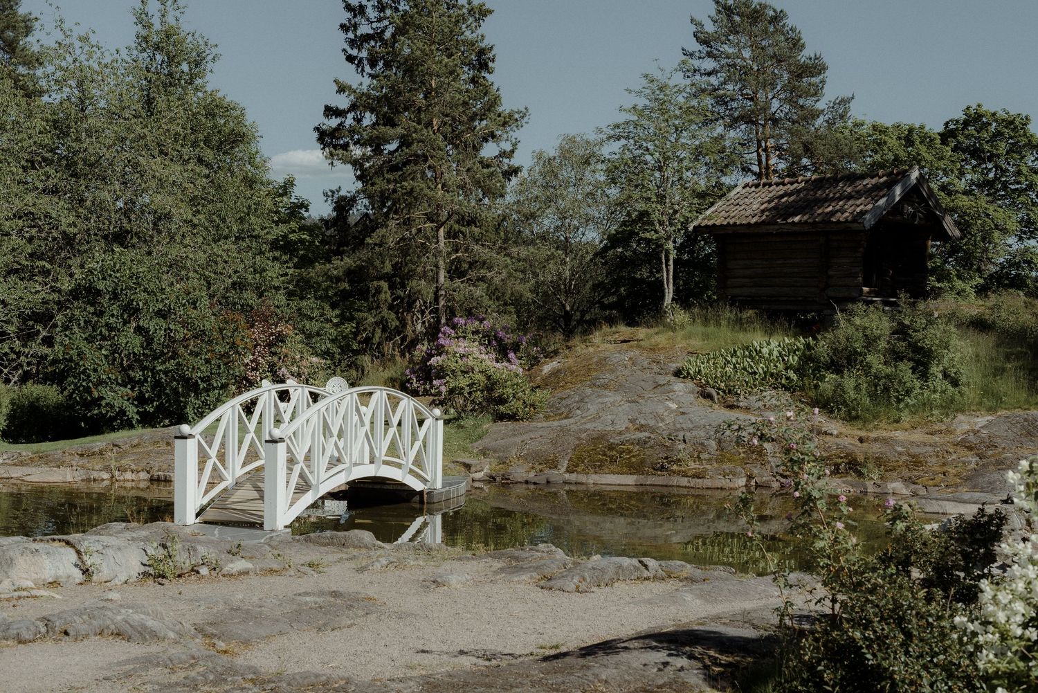A rustic white wooden bridge crosses over rocks near a small cabin in a Nordic summer landscape.