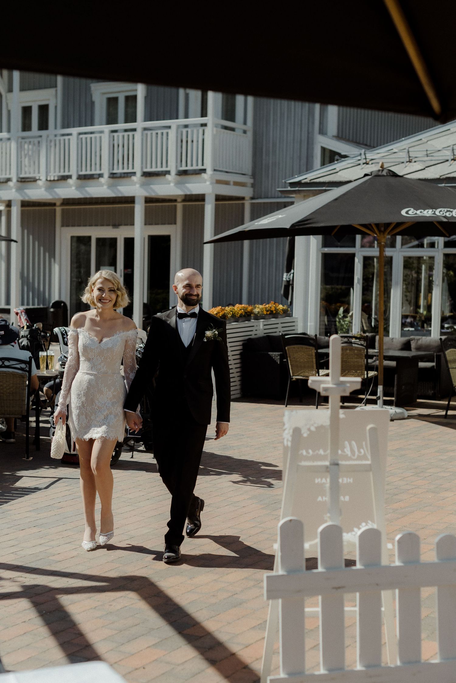 A pair walks along a sunny outdoor boardwalk with white railing details in a seaside setting.