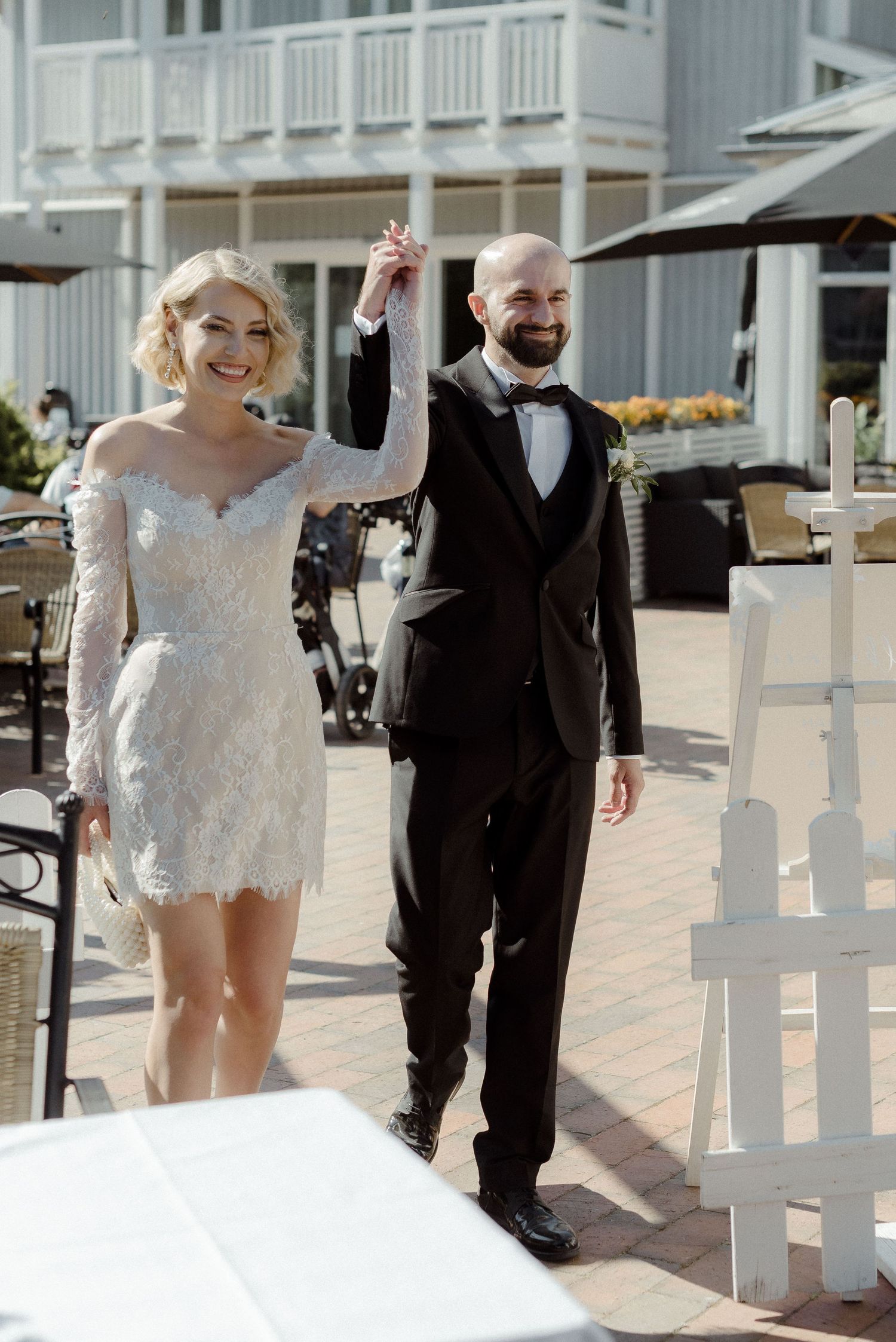A happy couple in wedding attire playfully walks and interacts along a white-railed outdoor walkway.
