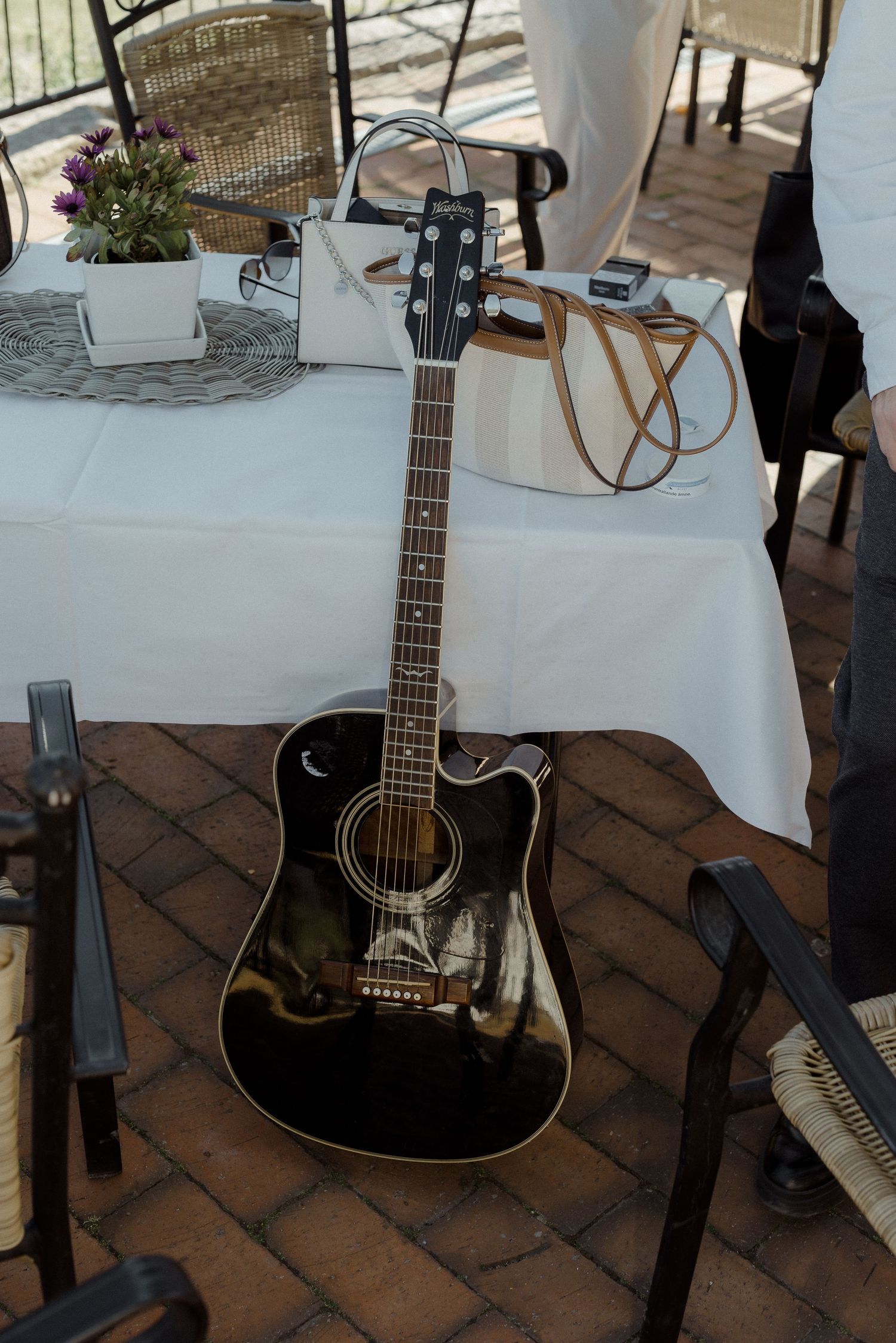 A black acoustic guitar rests on brick patio beside a white tablecloth setting with floral decor.
