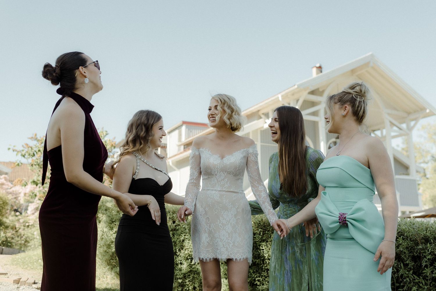 A group of women in formal dresses stand together laughing in a garden setting on a sunny day.