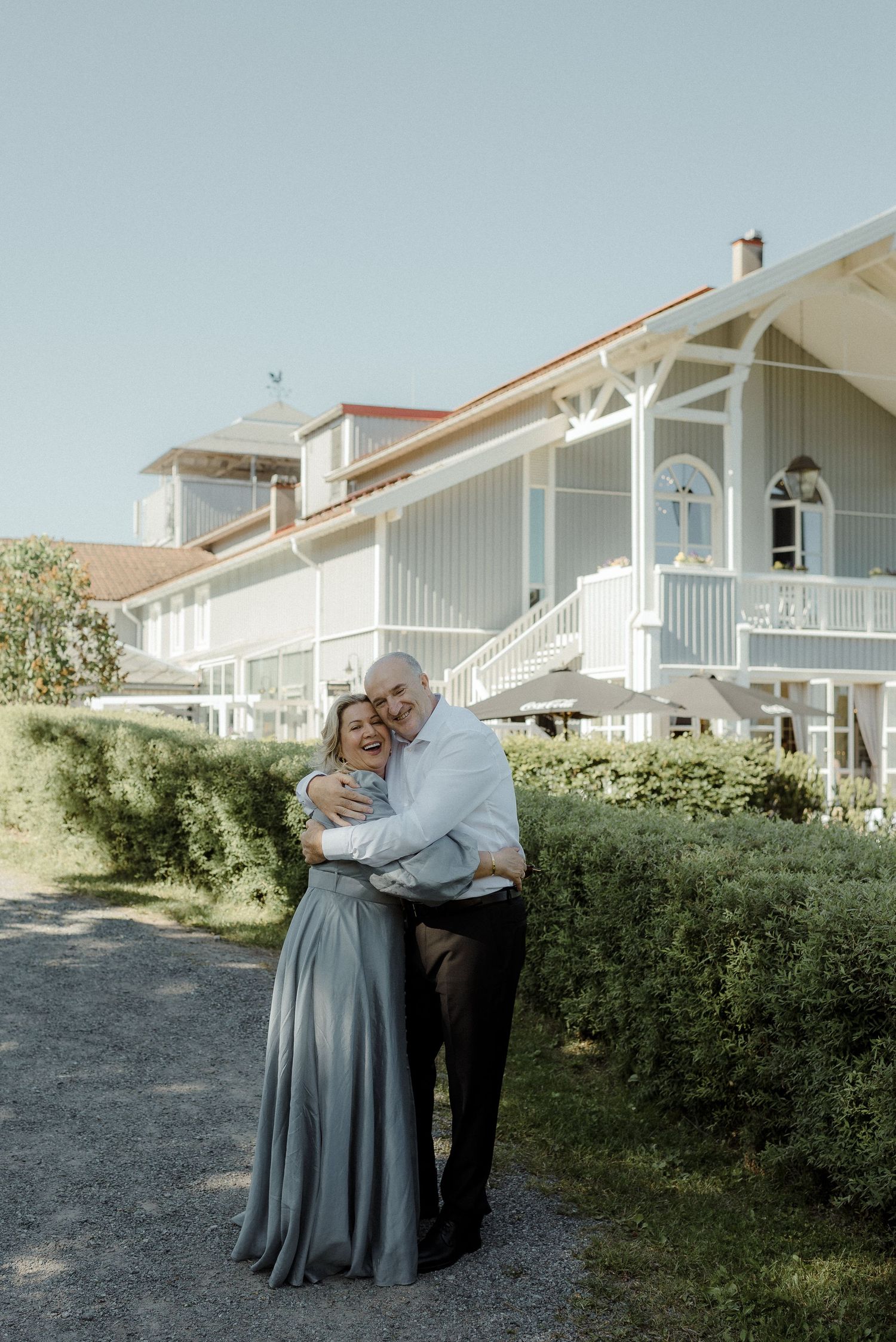 A couple embraces in front of a white coastal-style house with wraparound porch on a sunny summer day.