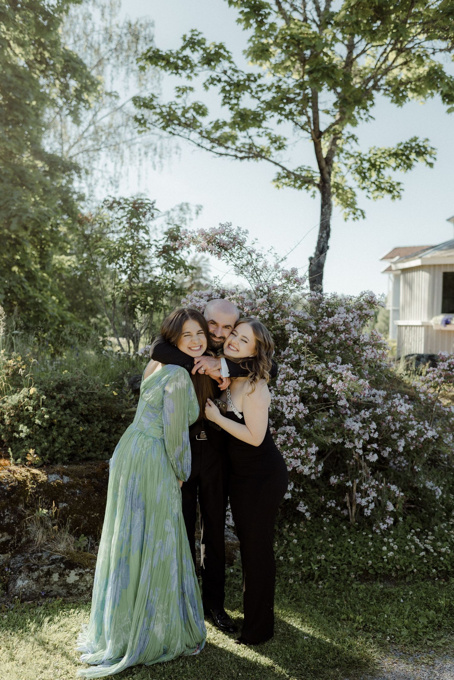 Three people embrace in a garden setting with blooming flowers and trees on a sunny day.