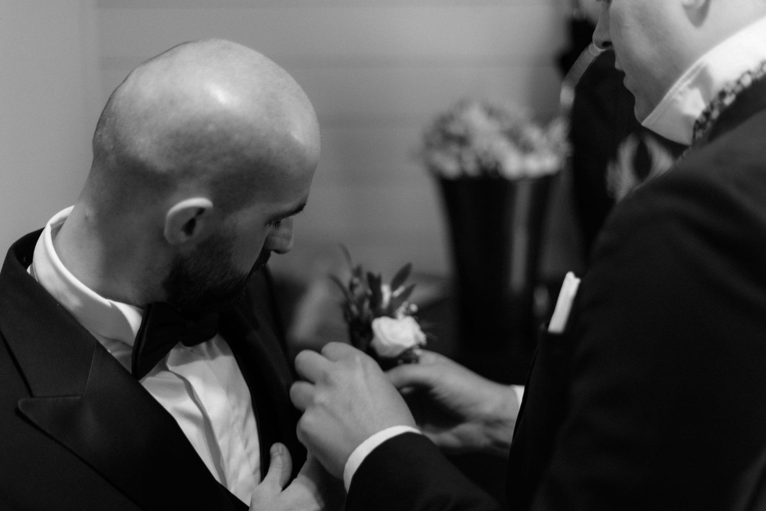Black and white photo showing someone helping to pin a boutonniere flower onto a groom's lapel before a wedding ceremony.