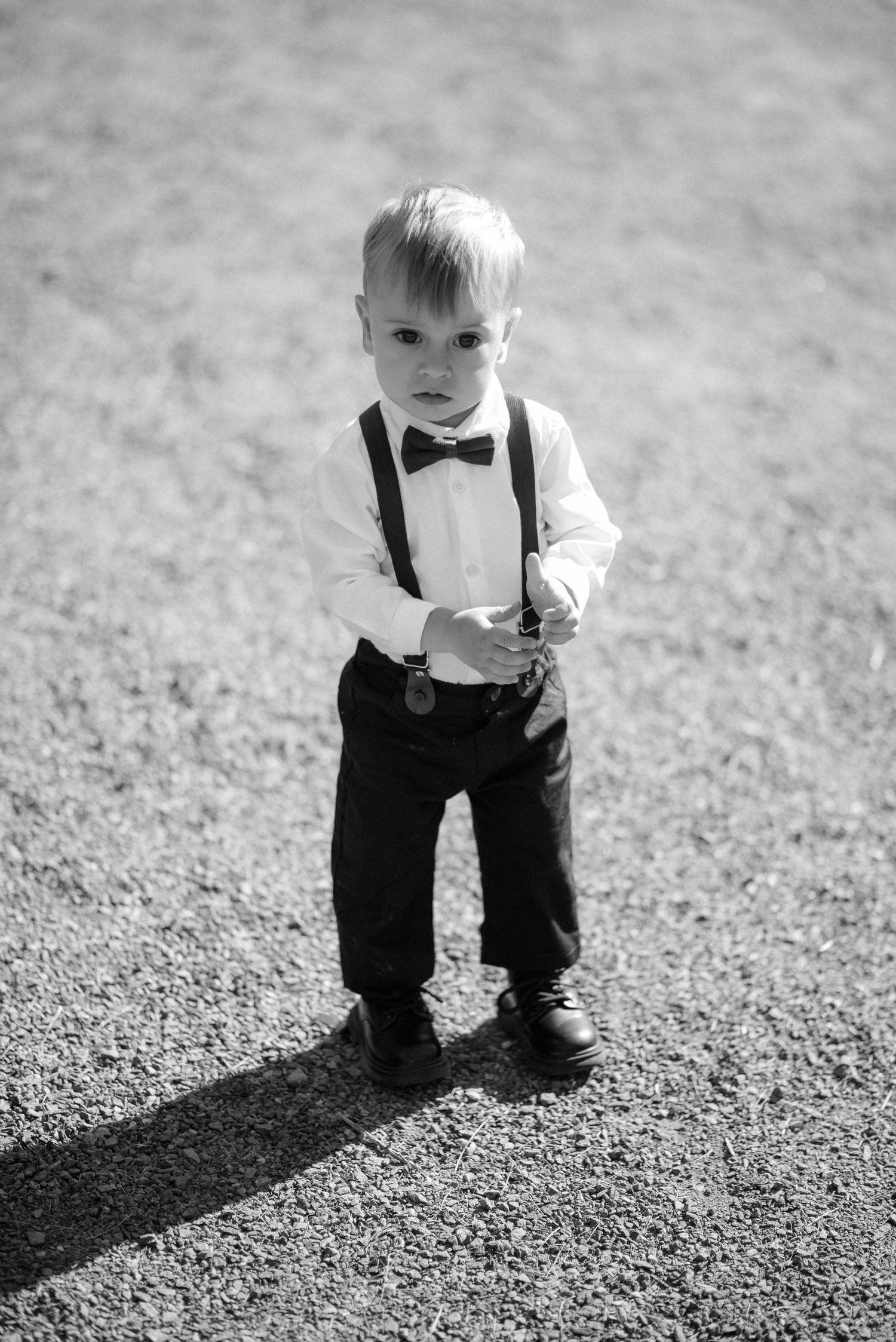 A small child wearing suspenders and bow tie stands confidently in formal attire in black and white photography.