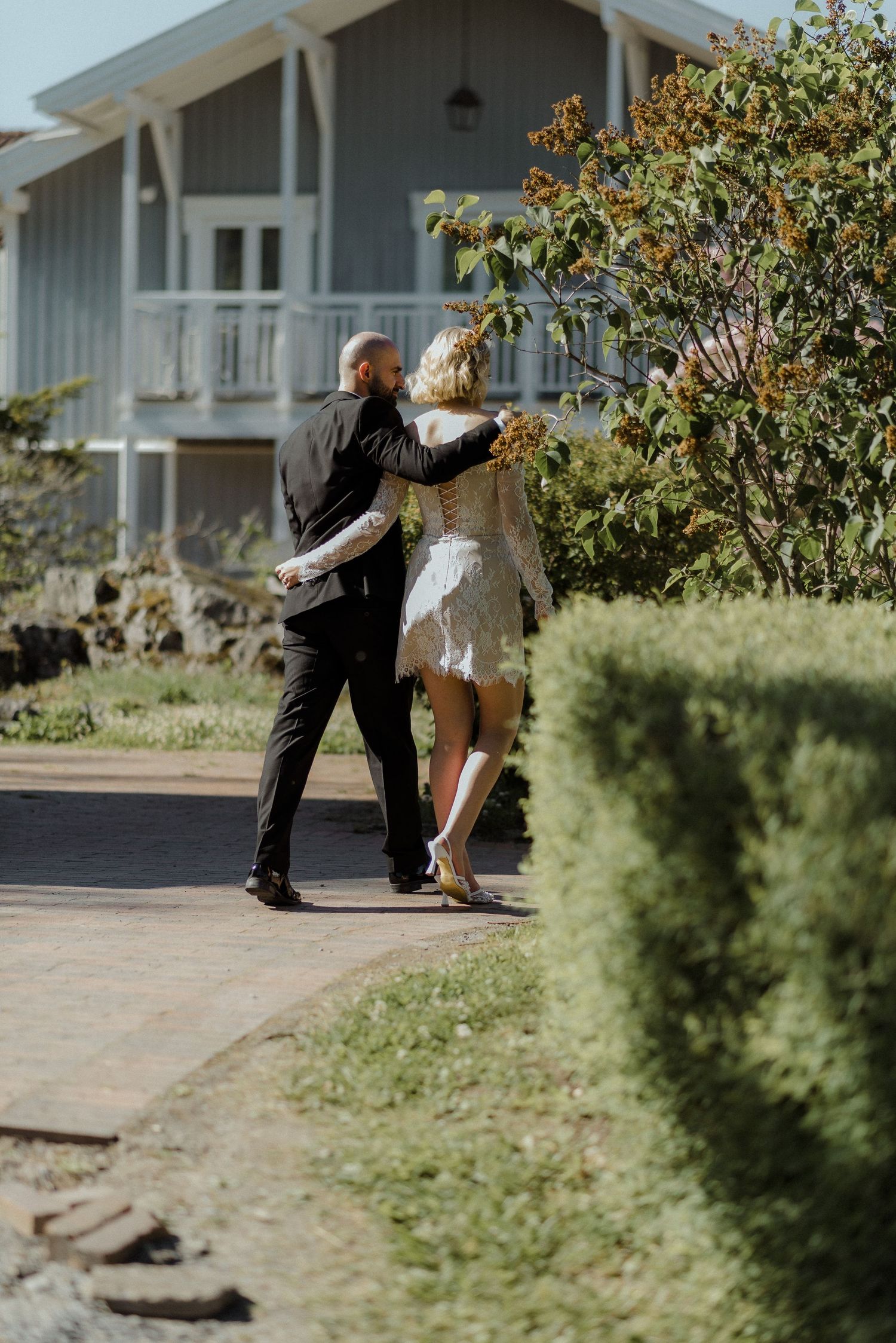 A couple dances outdoors on a paved path in front of a gray house with a white porch on a sunny day.