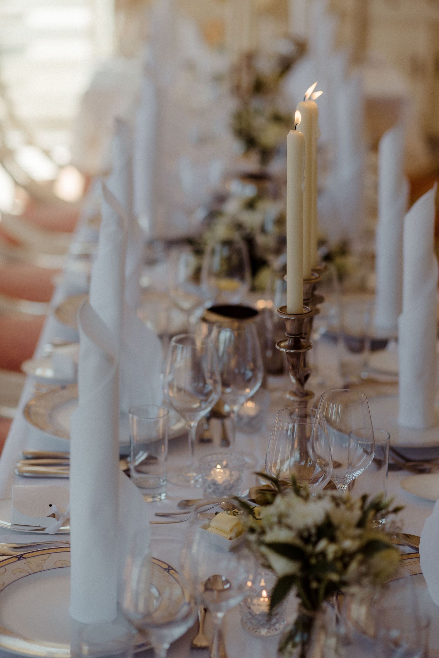 Elegant wedding table setting with white candles, wine glasses and floral arrangements on a white tablecloth.