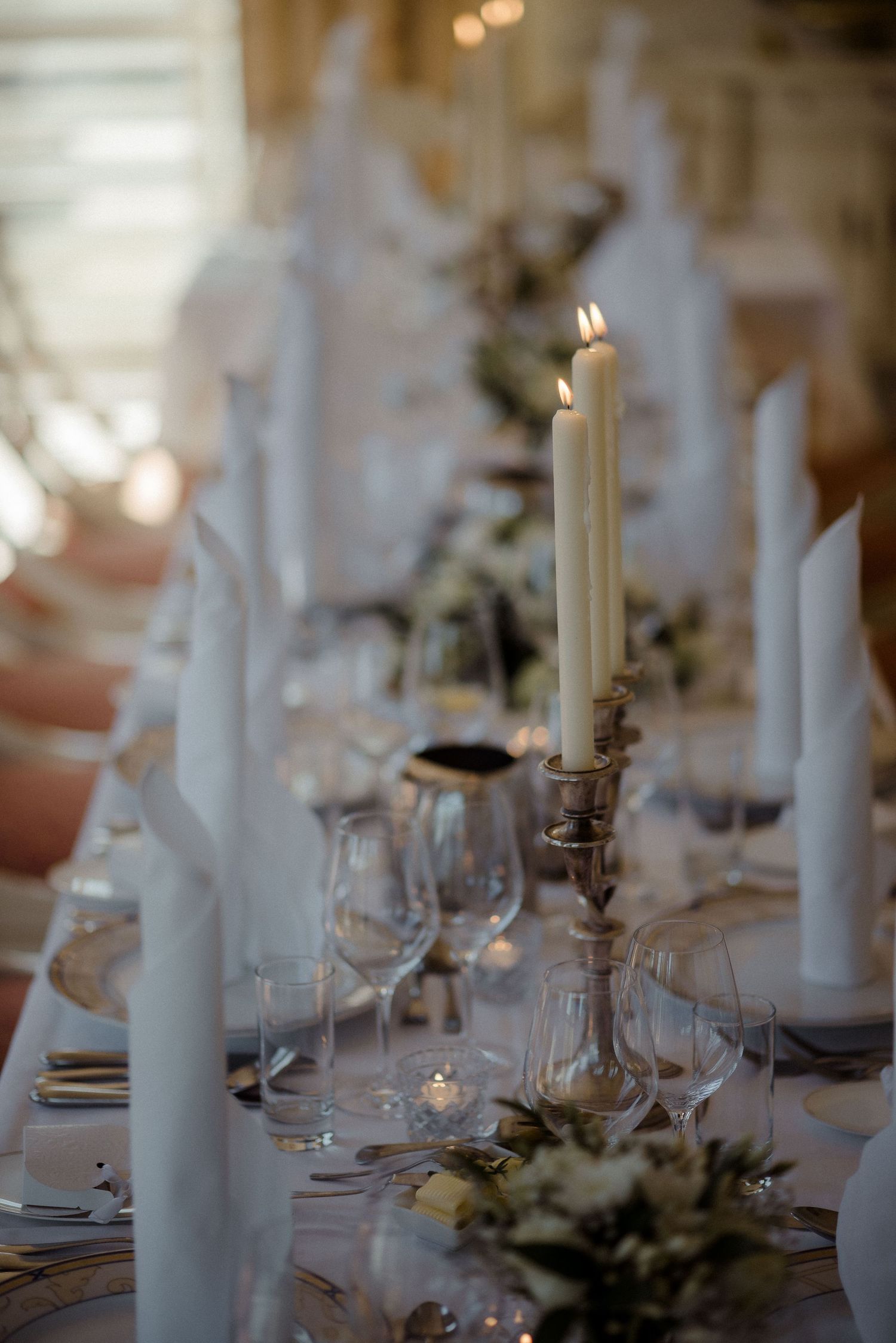 Elegant wedding table setting with tall white candles, wine glasses and floral centerpieces in soft lighting.