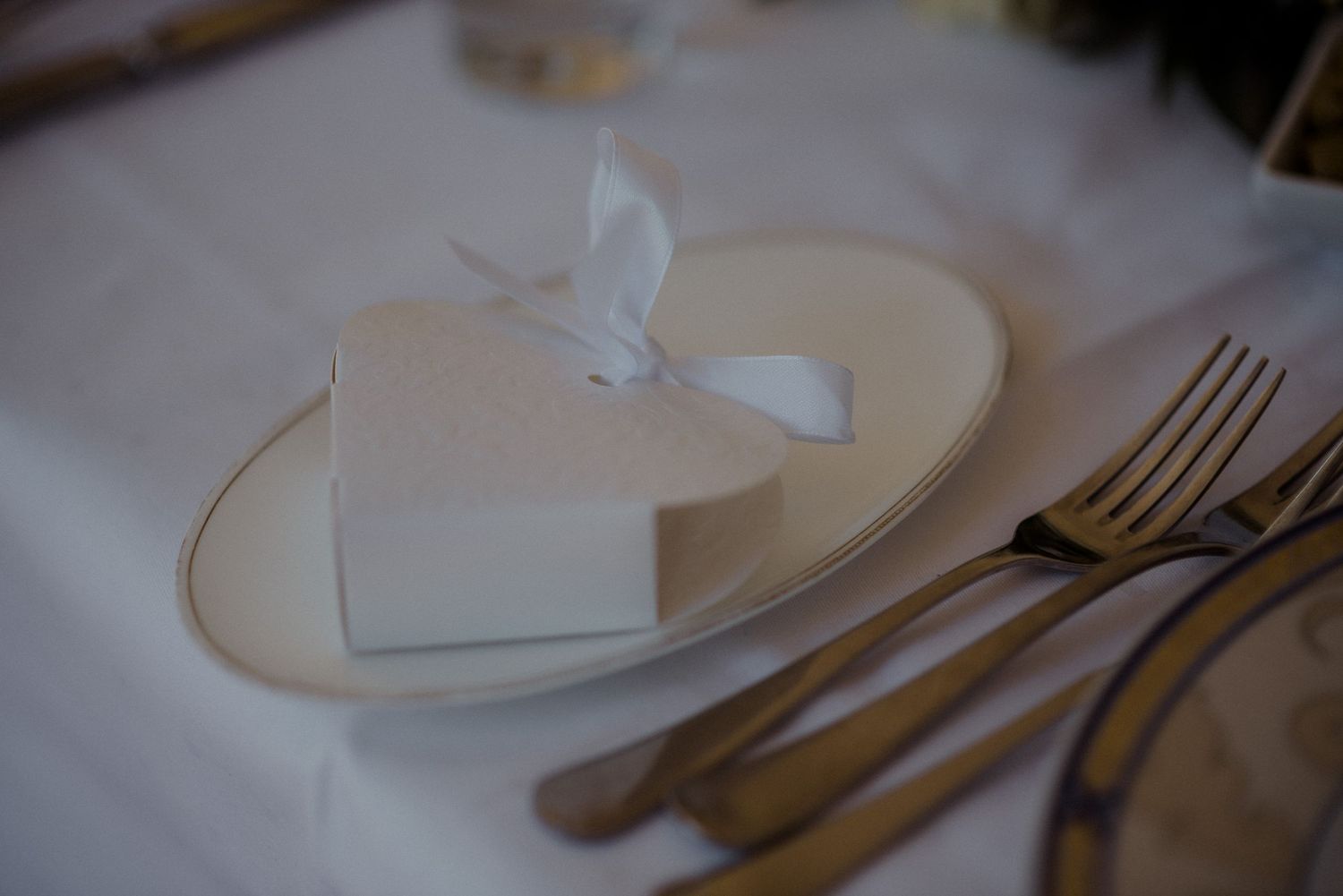 Heart-shaped favor box with white ribbon sits on elegant place setting with gold cutlery.
