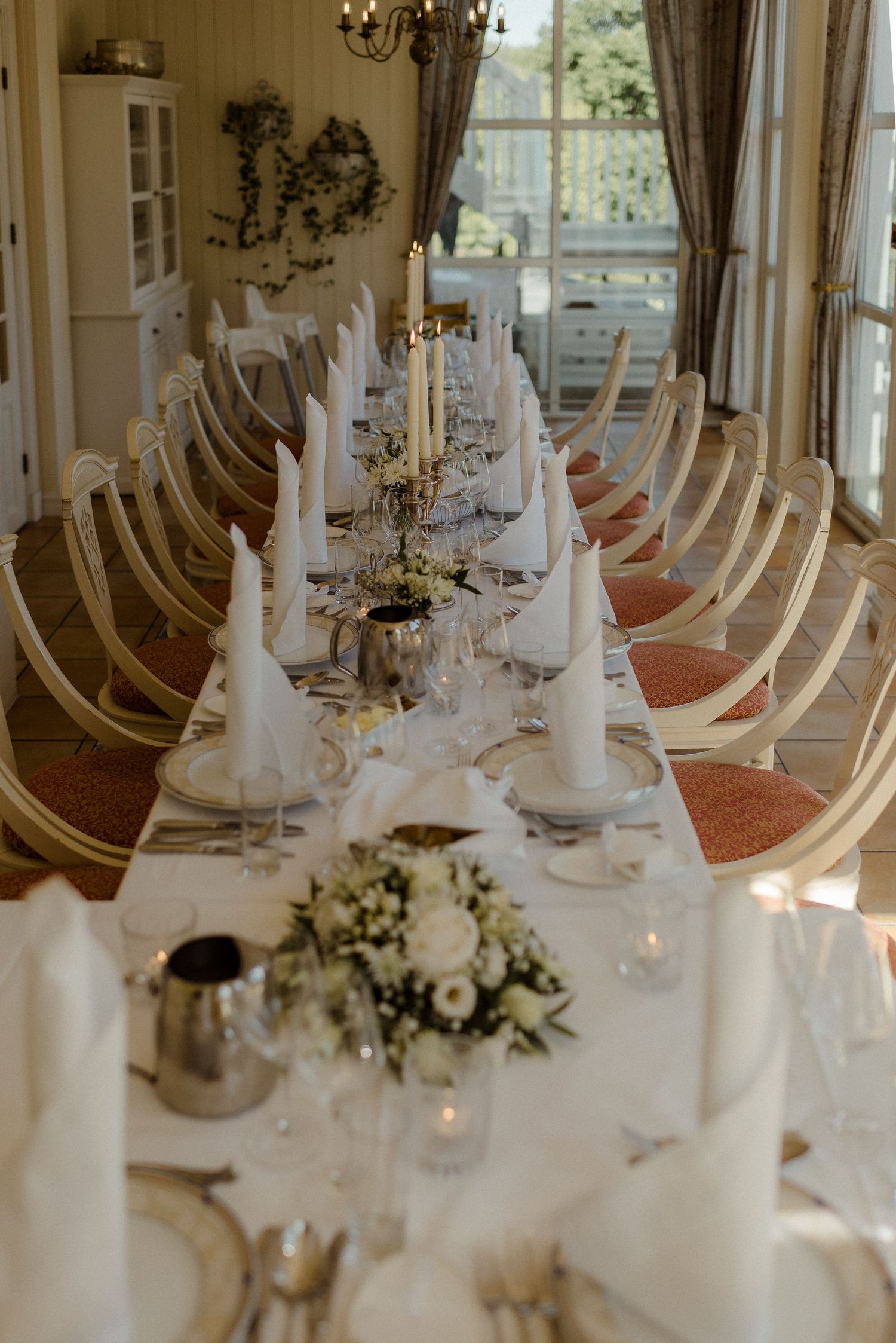 Elegant wedding reception table set with white linens, folded napkins and floral centerpieces in a bright sunlit room.