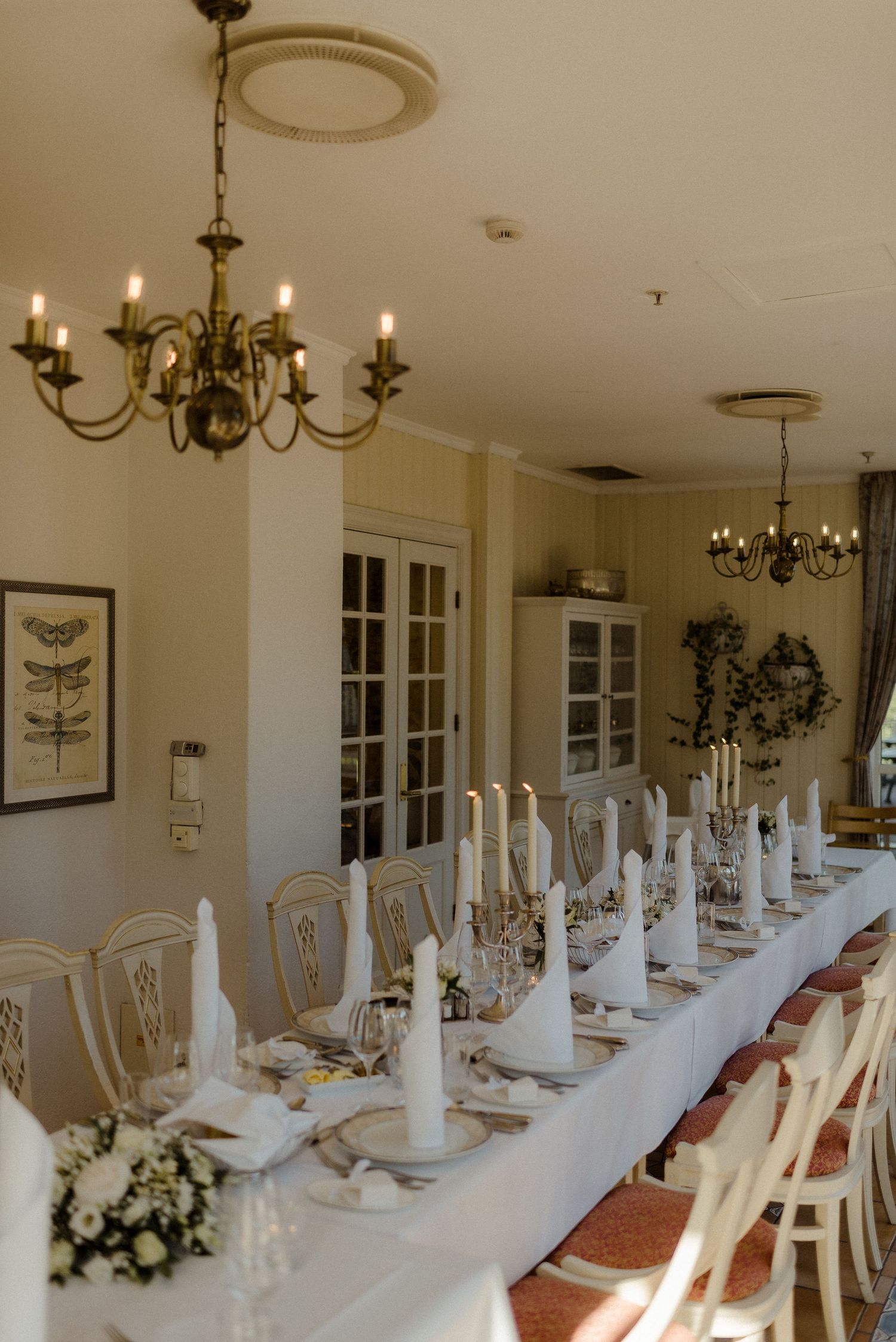 Elegant dining room setup with a long white table, traditional chandelier, and white chairs arranged for a formal event.