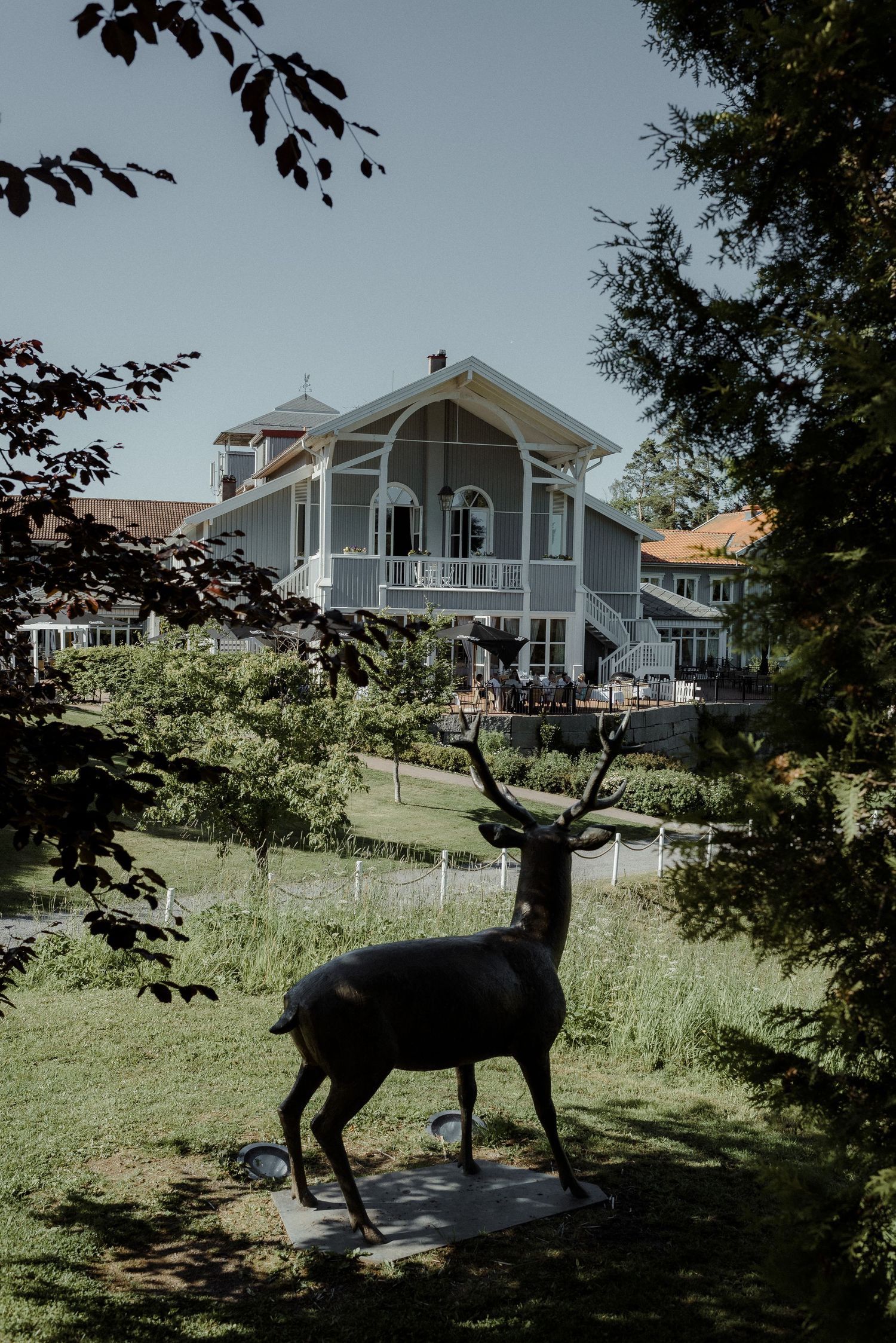 A large deer statue stands on a lawn in front of a white Victorian-style house with a wraparound porch.