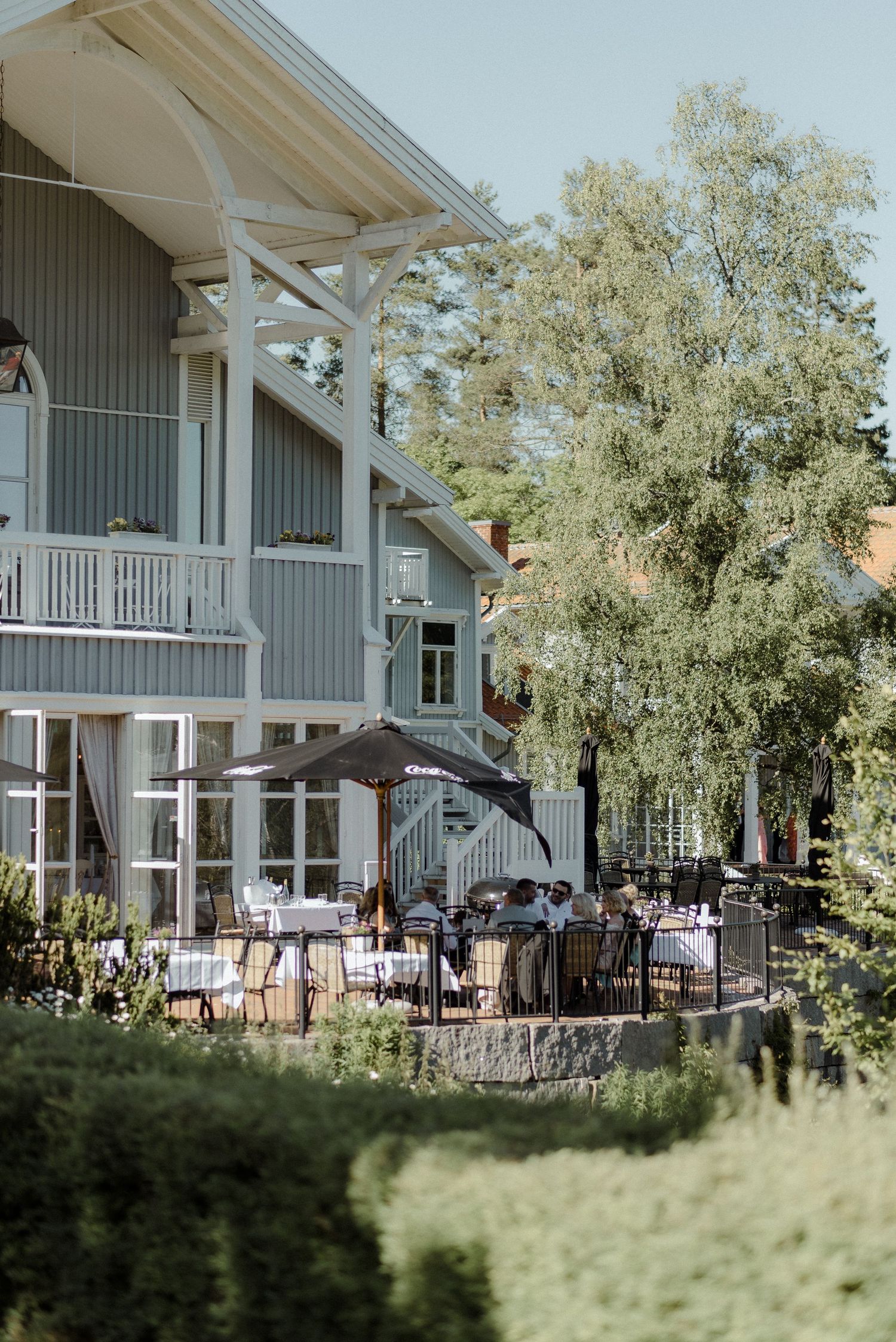 A charming gray house with white trim features an outdoor dining patio under tall trees with umbrellas and stone walls.