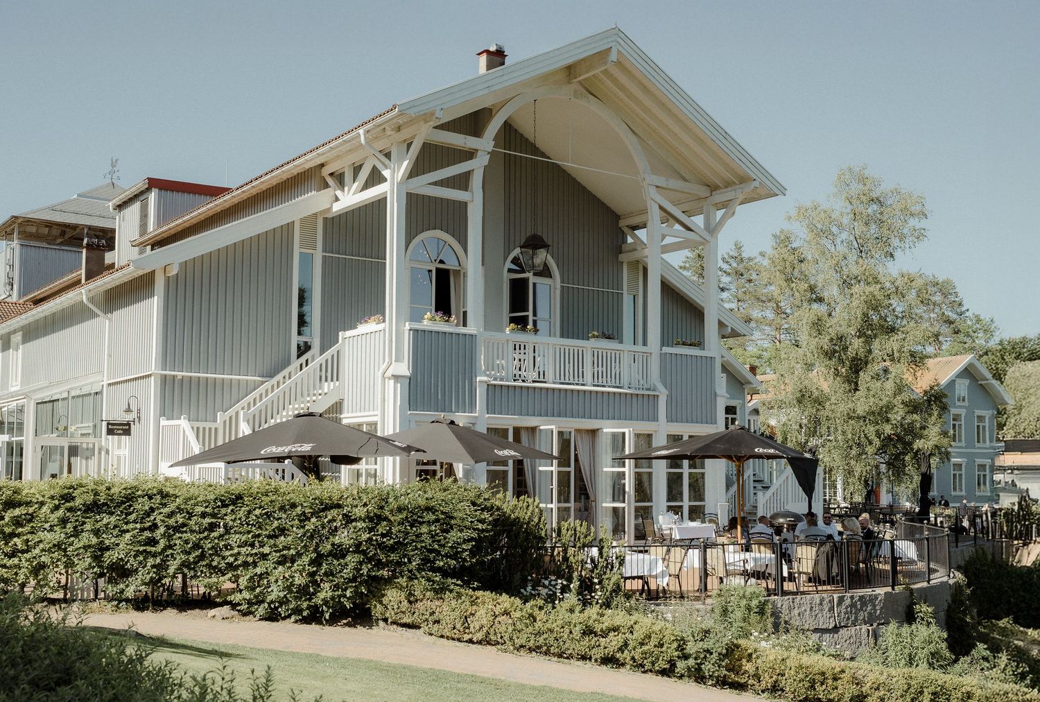 A charming gray Victorian-style house with wraparound porch and outdoor seating area surrounded by manicured hedges.