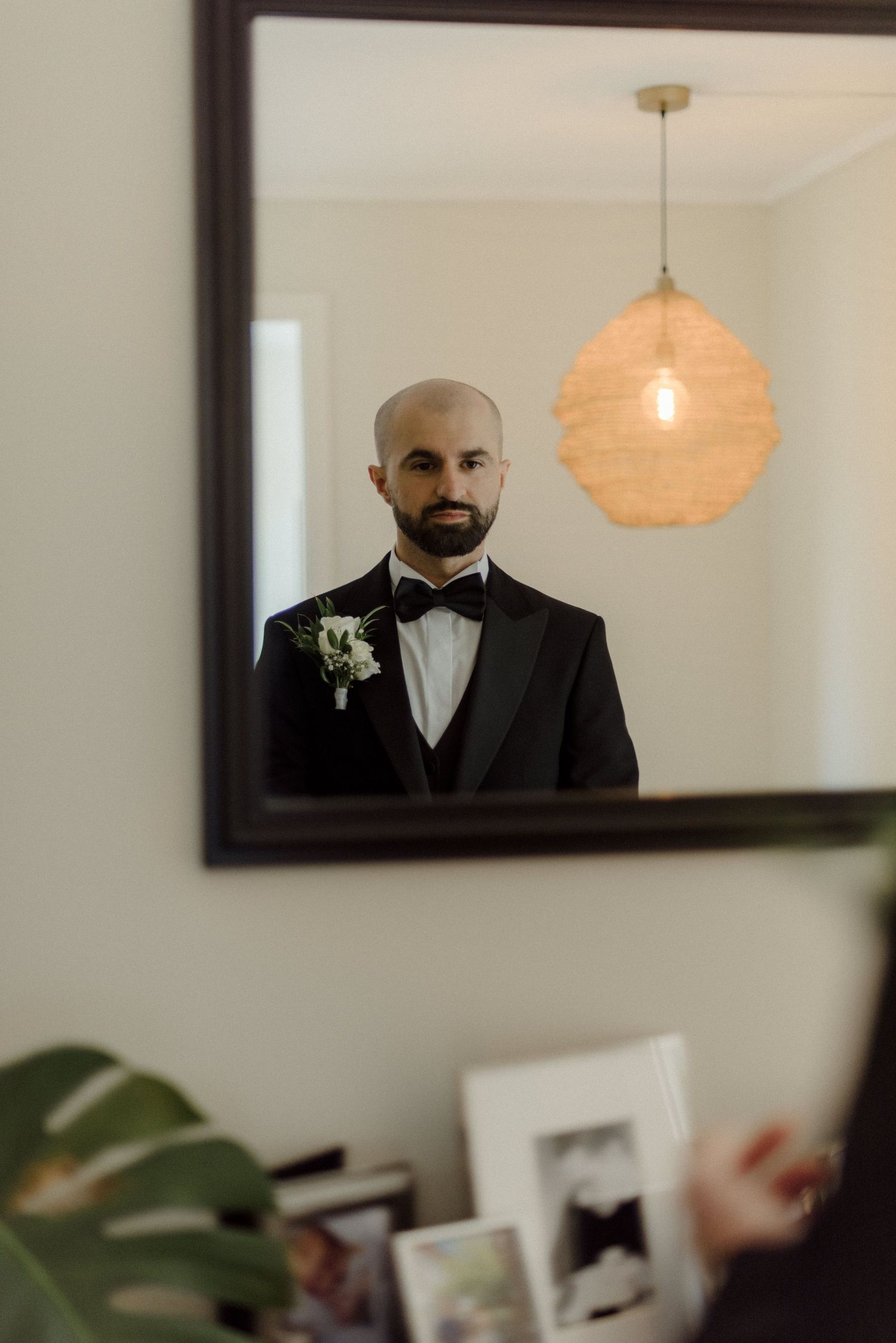 Mirror reflection showing a formal wedding attire with black tuxedo and white boutonniere under a warm pendant light.