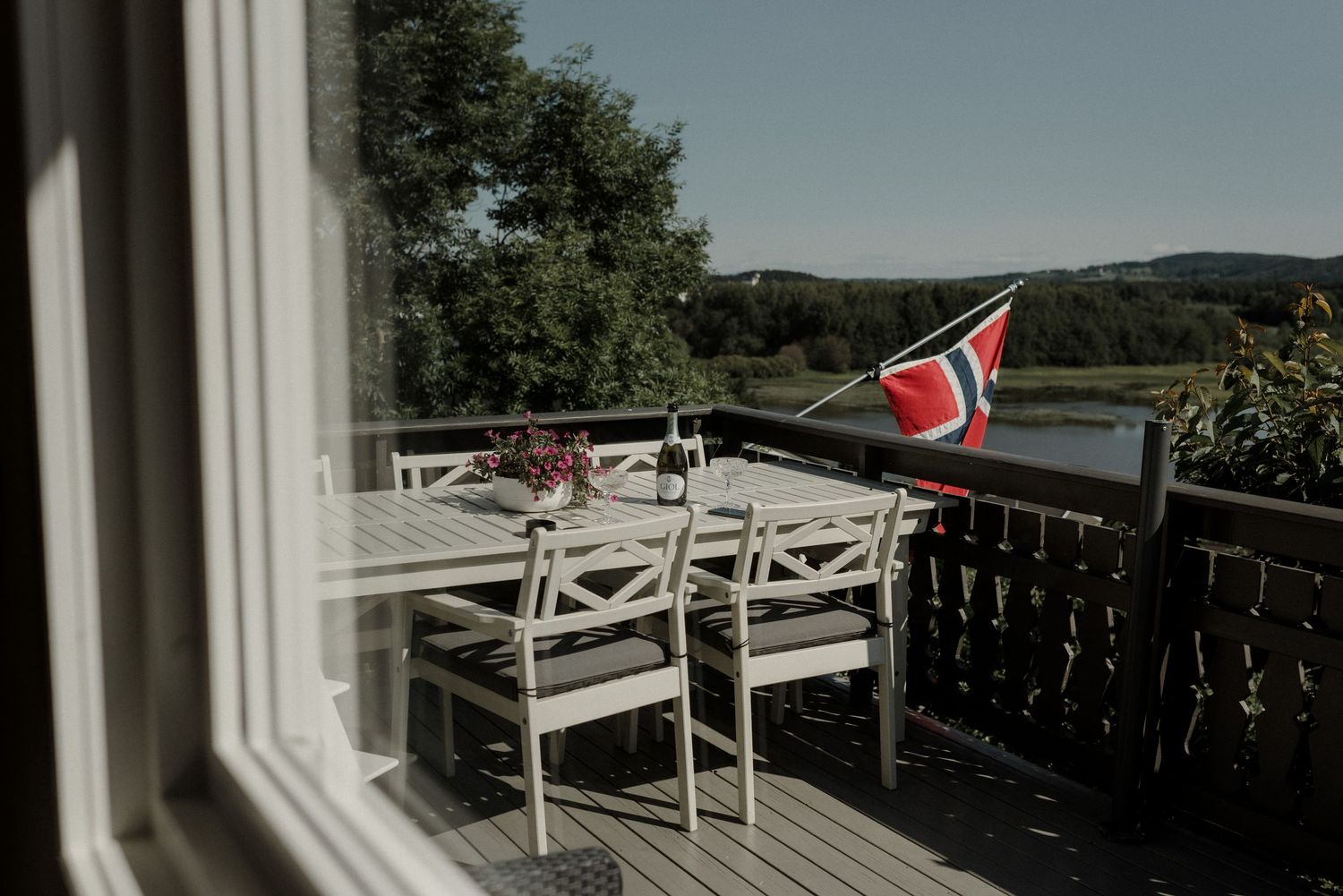 Norwegian flag waves on a wooden deck patio with white outdoor furniture overlooking scenic countryside views.