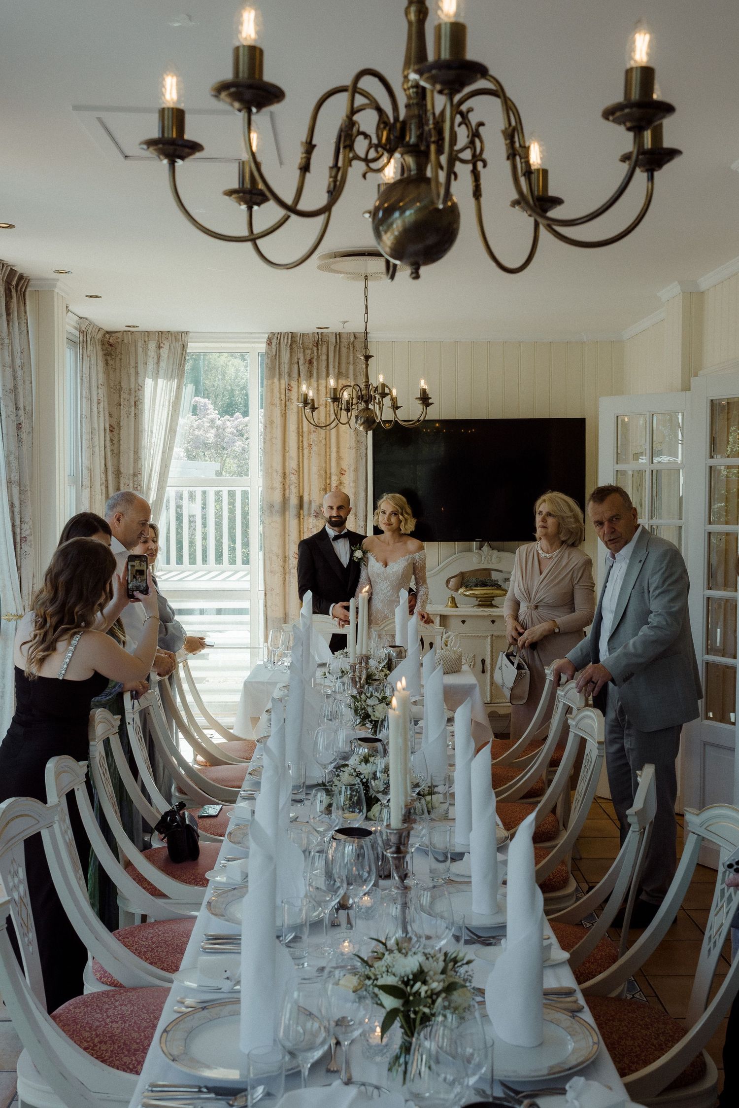 Elegant wedding reception table set with white linens, candles and floral centerpieces under brass chandeliers in bright room.