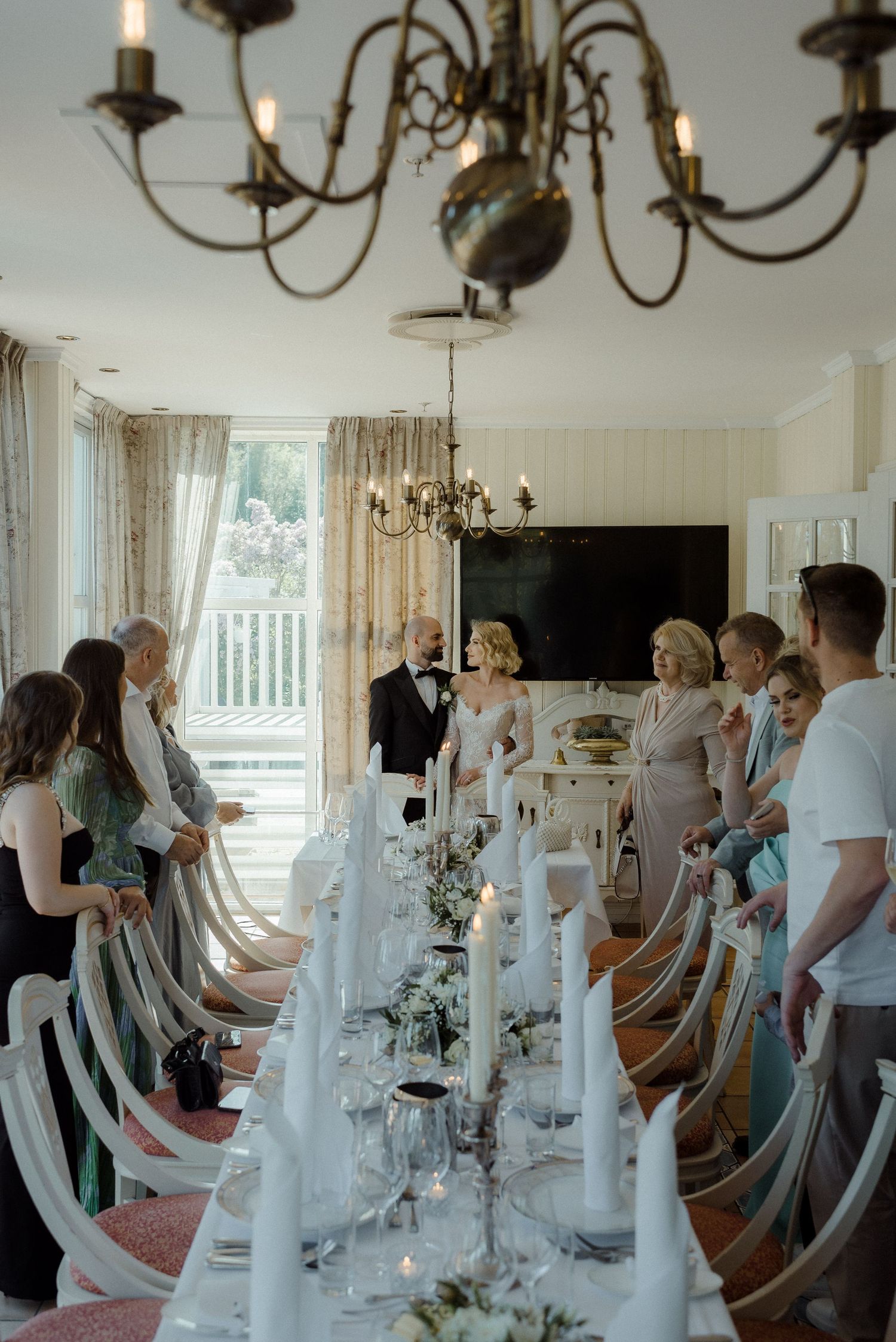Elegant wedding reception table setting with white candles and floral decorations under vintage chandelier in bright indoor space.