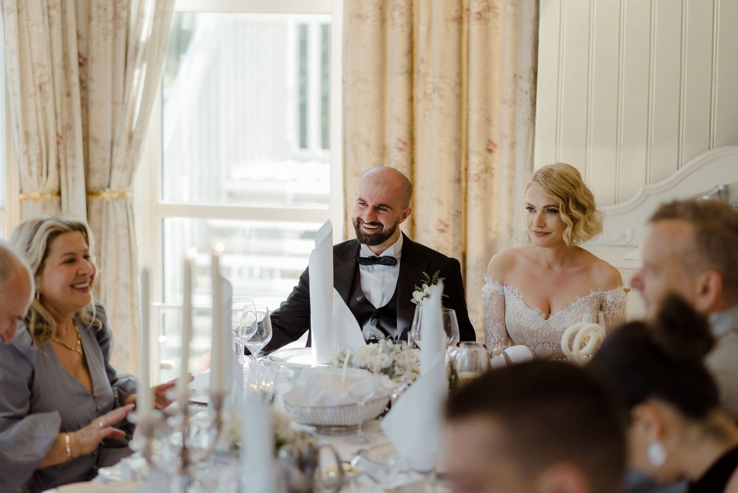 Group getting ready in a bridal suite with champagne and formal attire in a warm sunlit room filled with laughter.