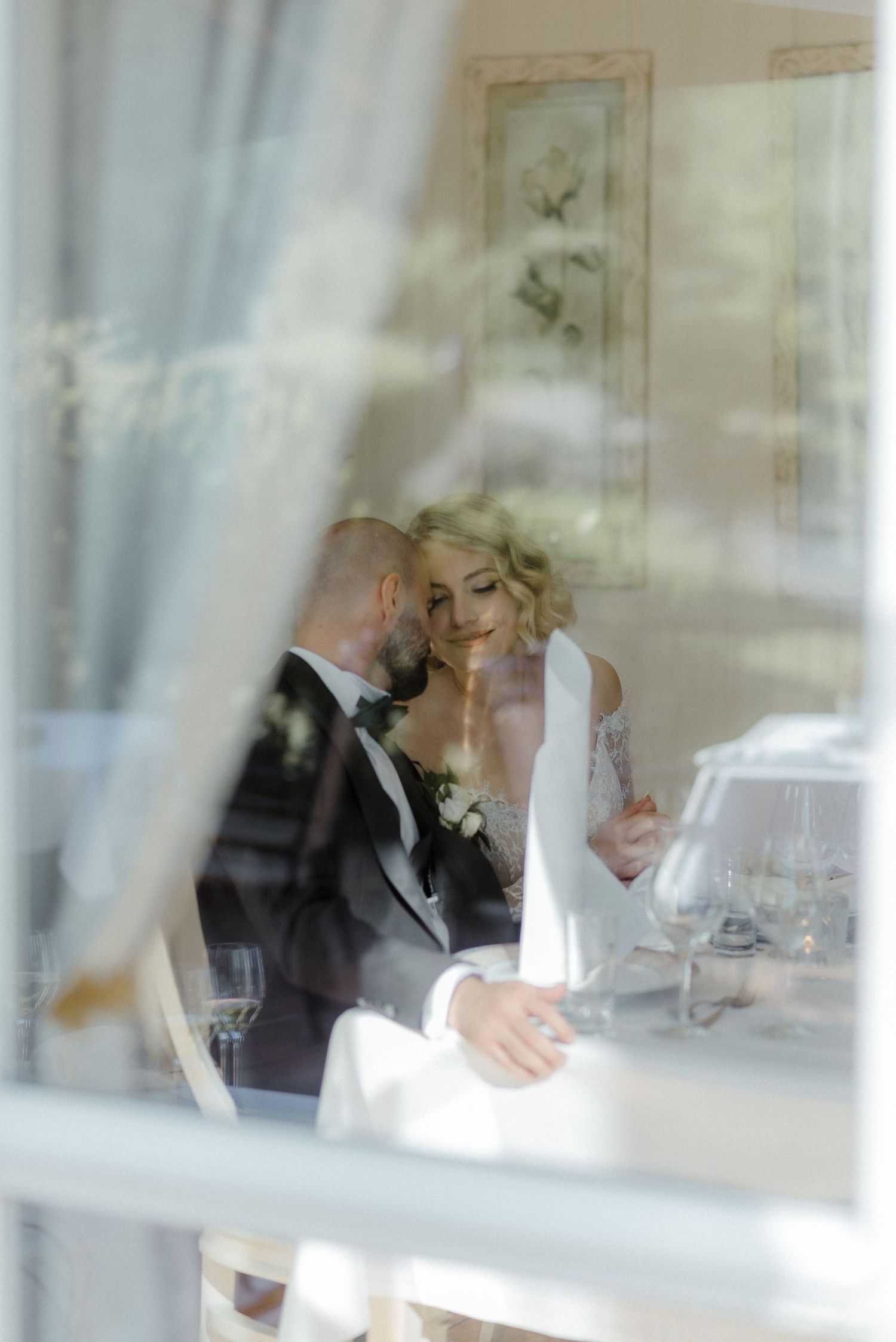 Wedding reception guests seated at elegant tables in a bright sunlit room with white decor and glassware.