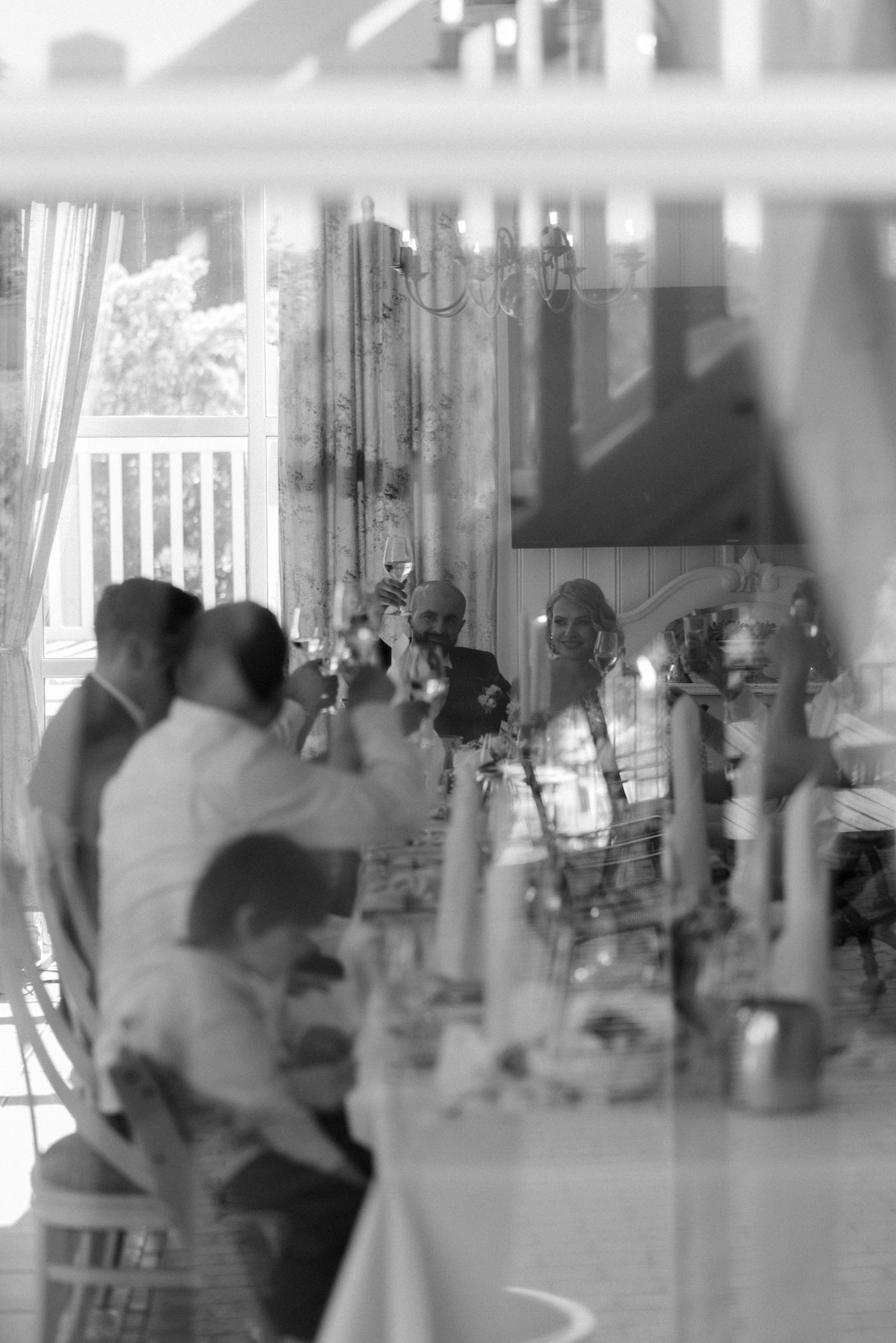 A black and white photograph showing people seated at a dining table through sheer curtains in soft focus.