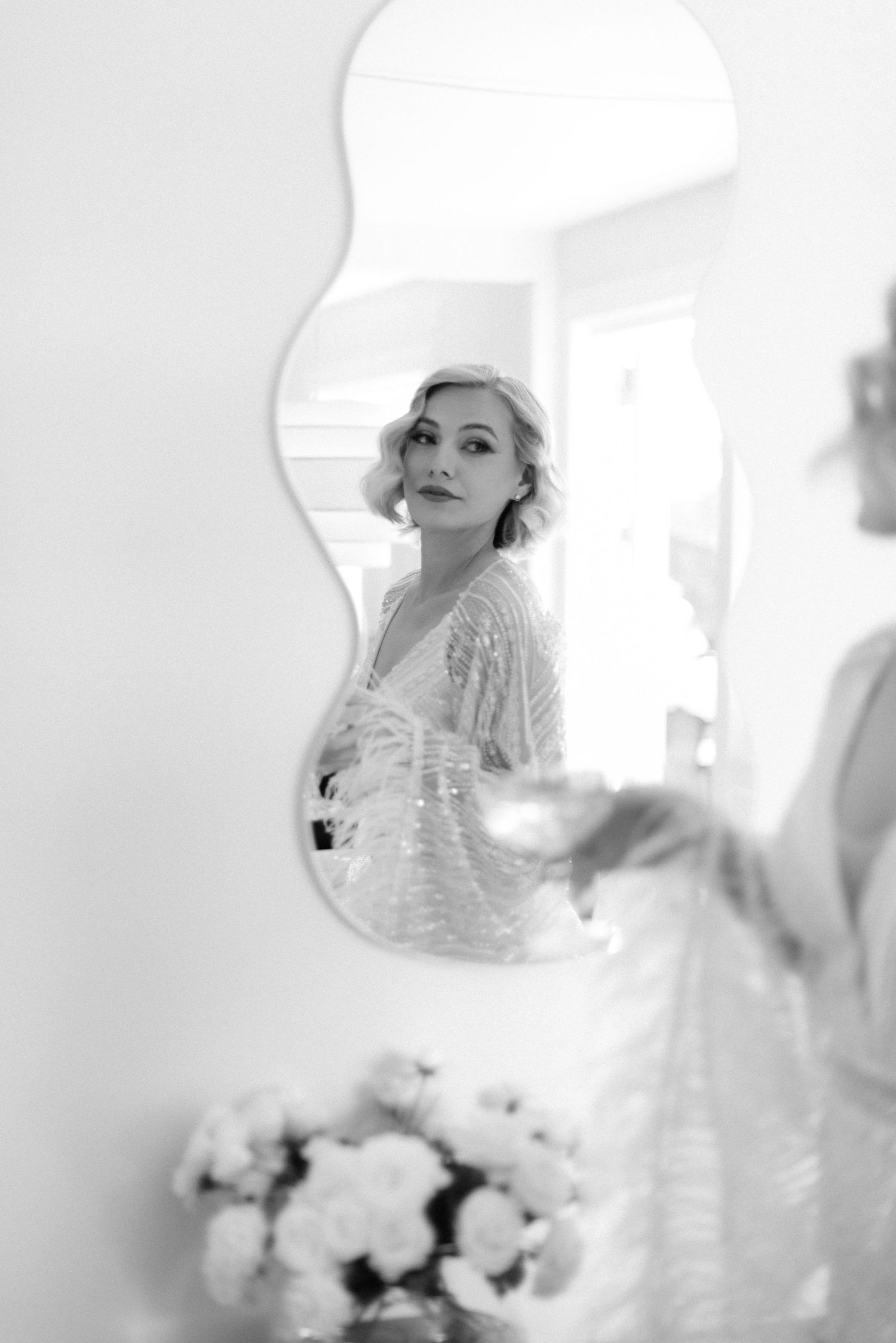 Artistic black and white reflection of a bride in an ornate mirror with flowers in the foreground.