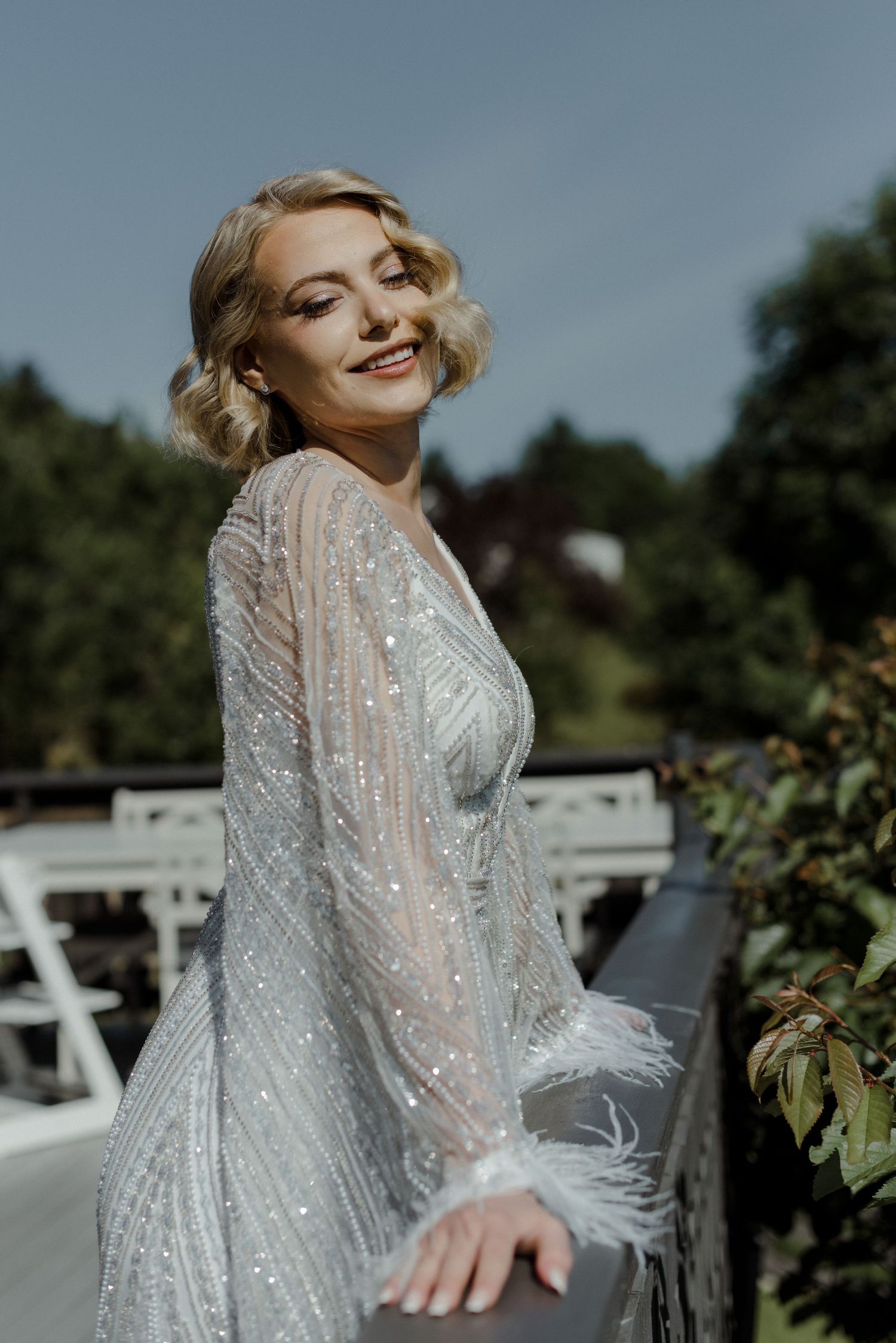 A beaming person in a shimmering long-sleeved wedding gown with sequins poses outdoors on a sunny day.