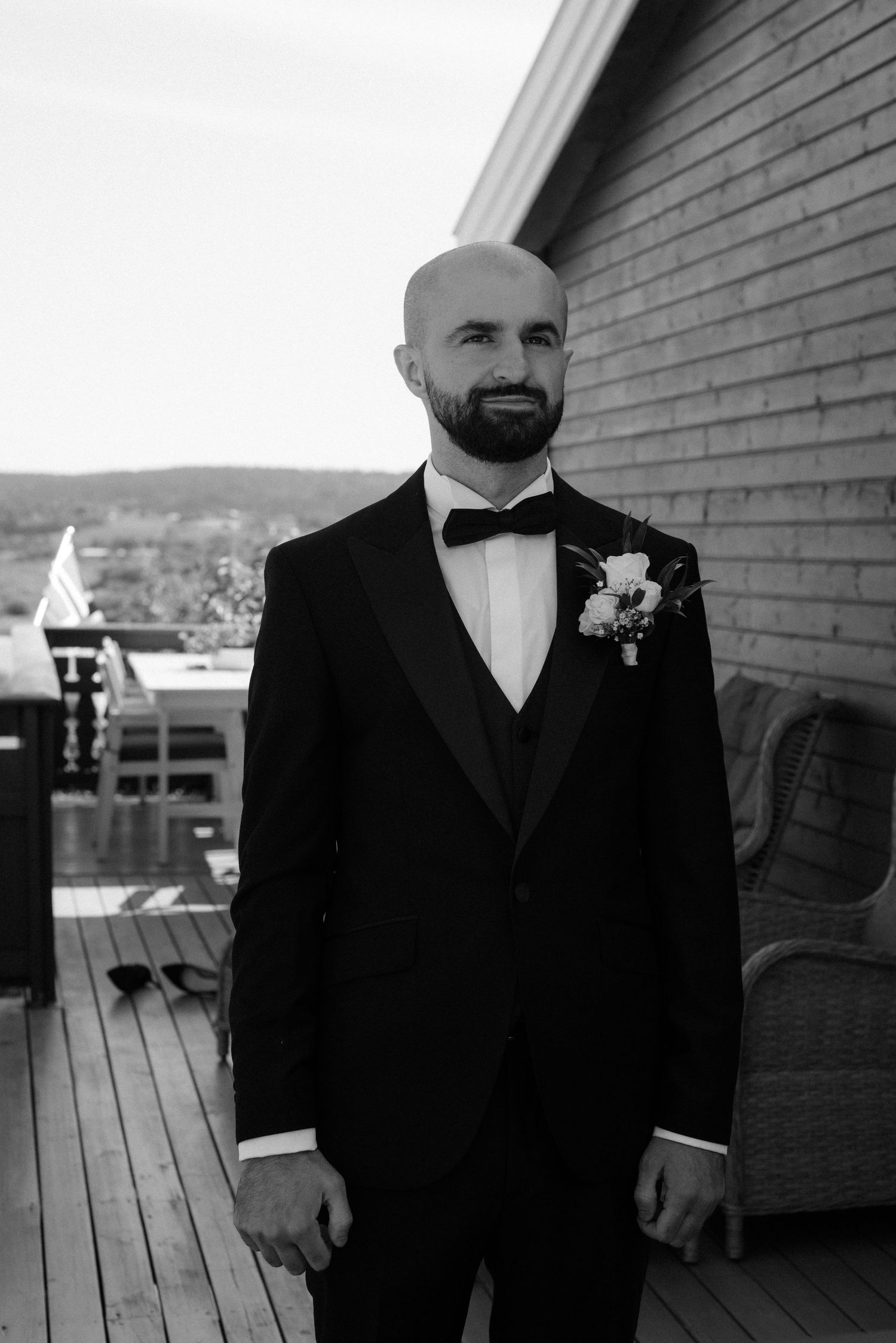 A black and white portrait of a groom in a classic tuxedo with bow tie and boutonniere standing on a wooden deck.