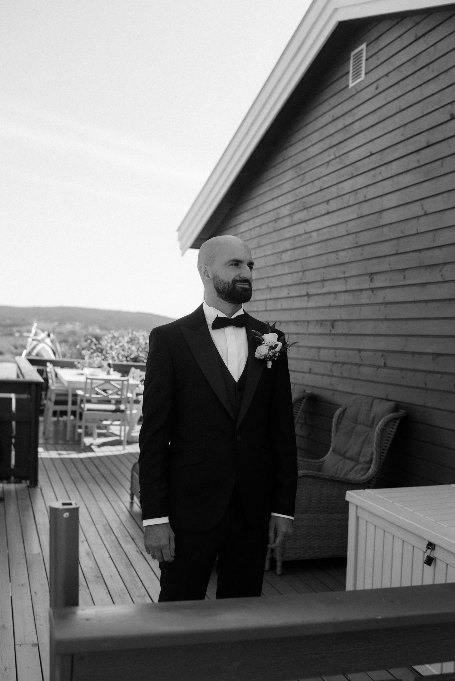 A black and white photo of someone in a tuxedo with a bow tie and boutonniere standing on a deck by a house.