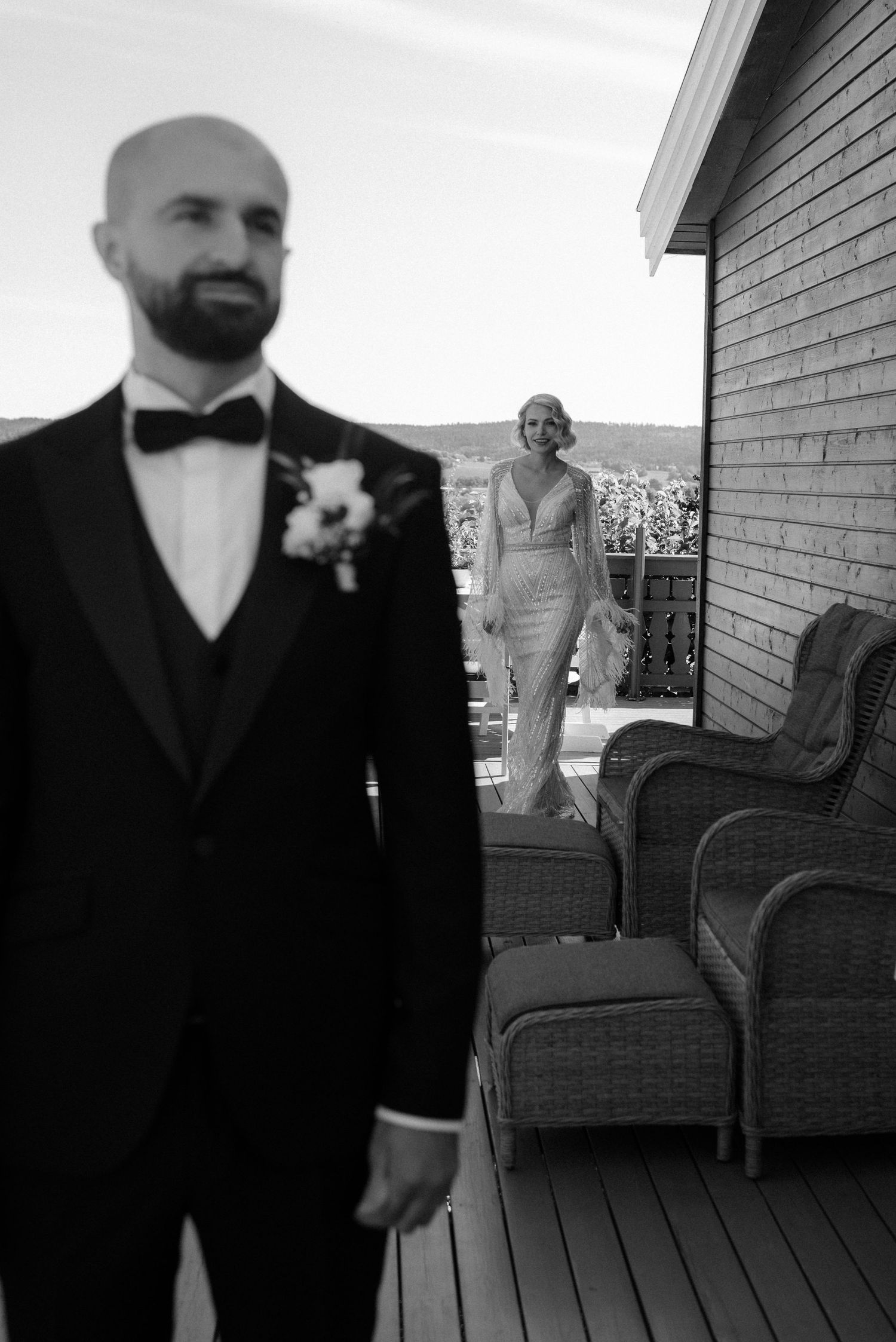 Black and white photo of people in formal attire on a covered deck with a brick wall and dark chairs.