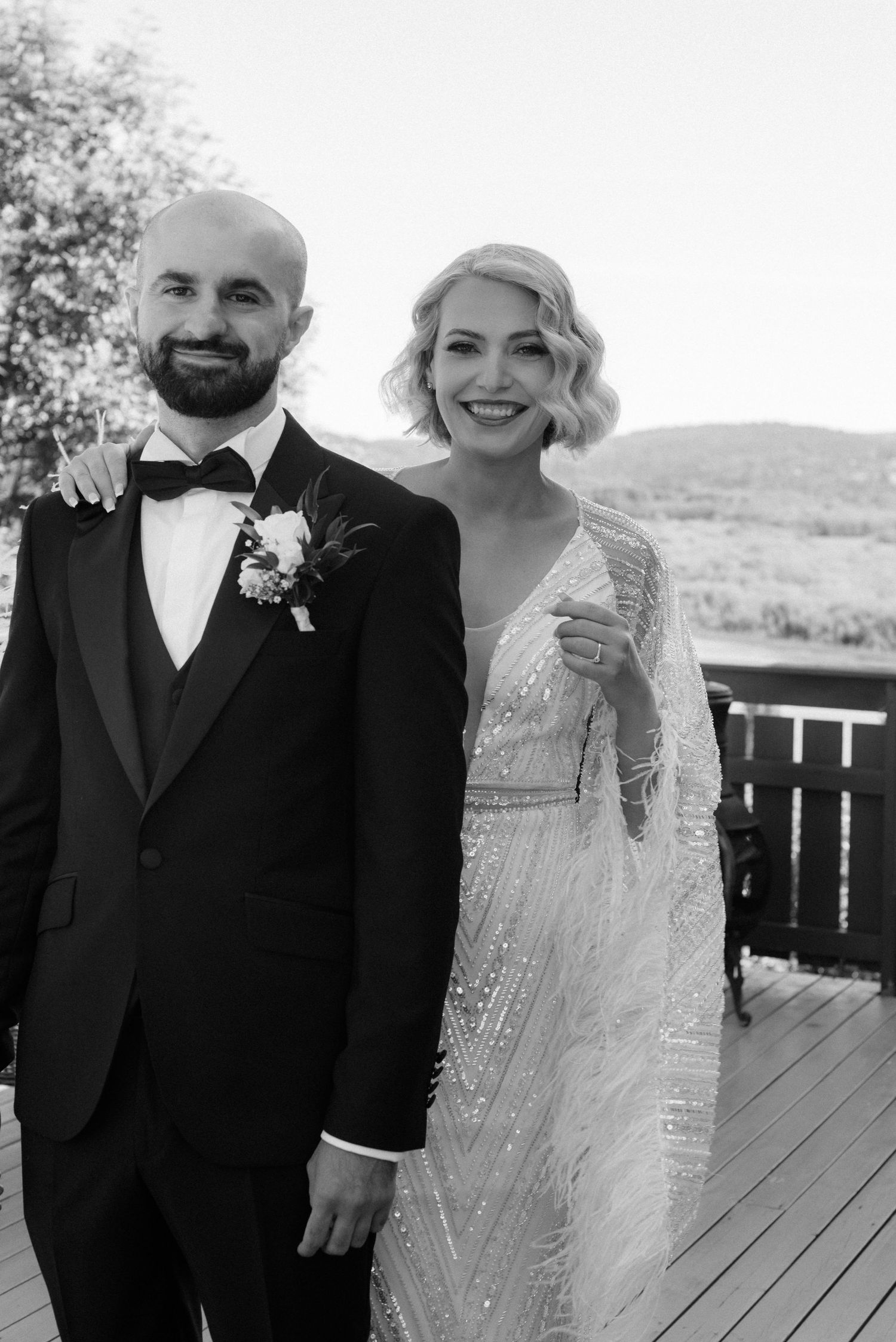 A black and white wedding photo of a couple on a deck overlooking mountains, with the bride in a beaded gown.