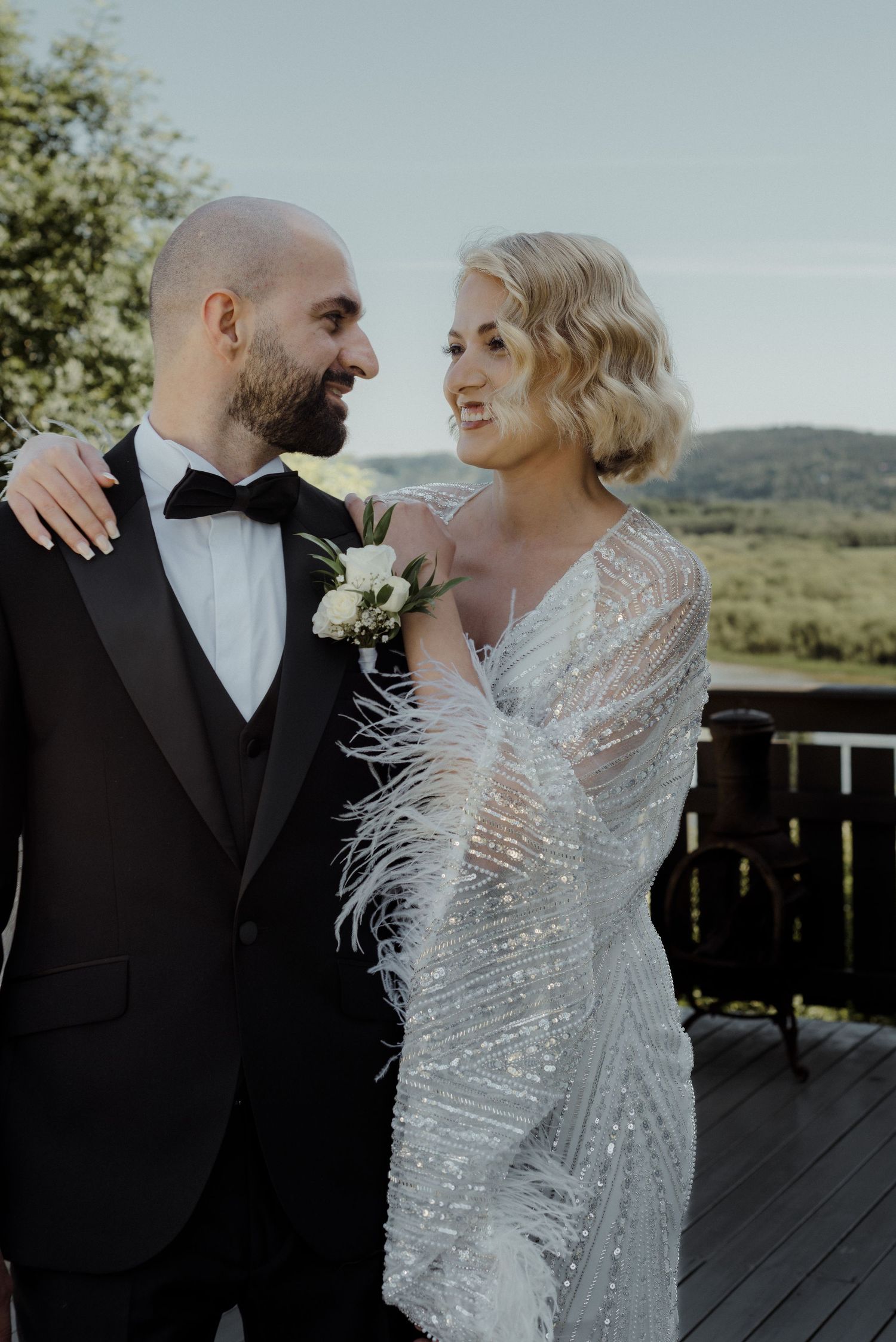 A bride in a feathered sequin gown and groom in black tuxedo share a joyful moment on an outdoor deck.