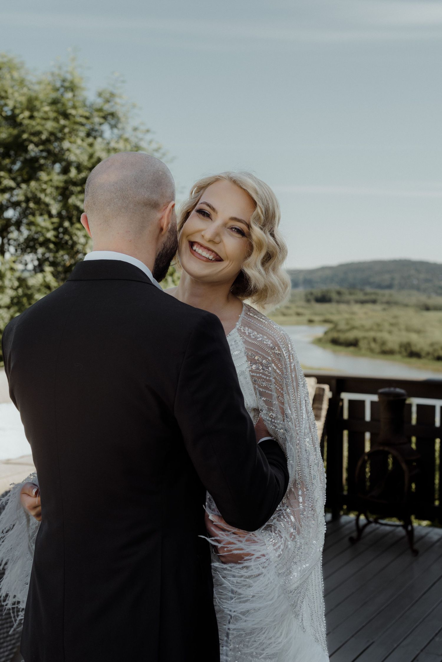 Newlywed couple embracing on outdoor deck with scenic mountain backdrop and natural lighting.