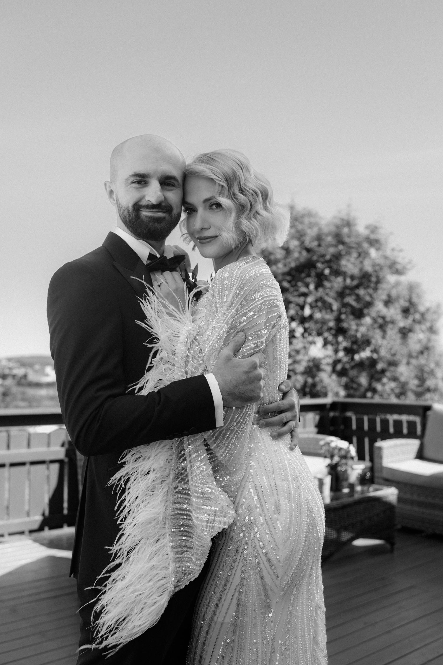 Black and white portrait of couple in elegant wedding attire with bride wearing feathered gown on outdoor terrace.