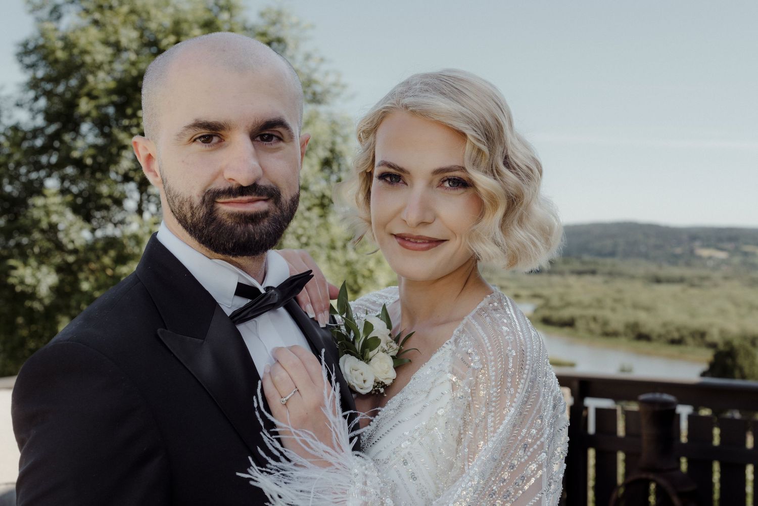 A wedding couple pose together outdoors on a sunny day with green hills in the background.