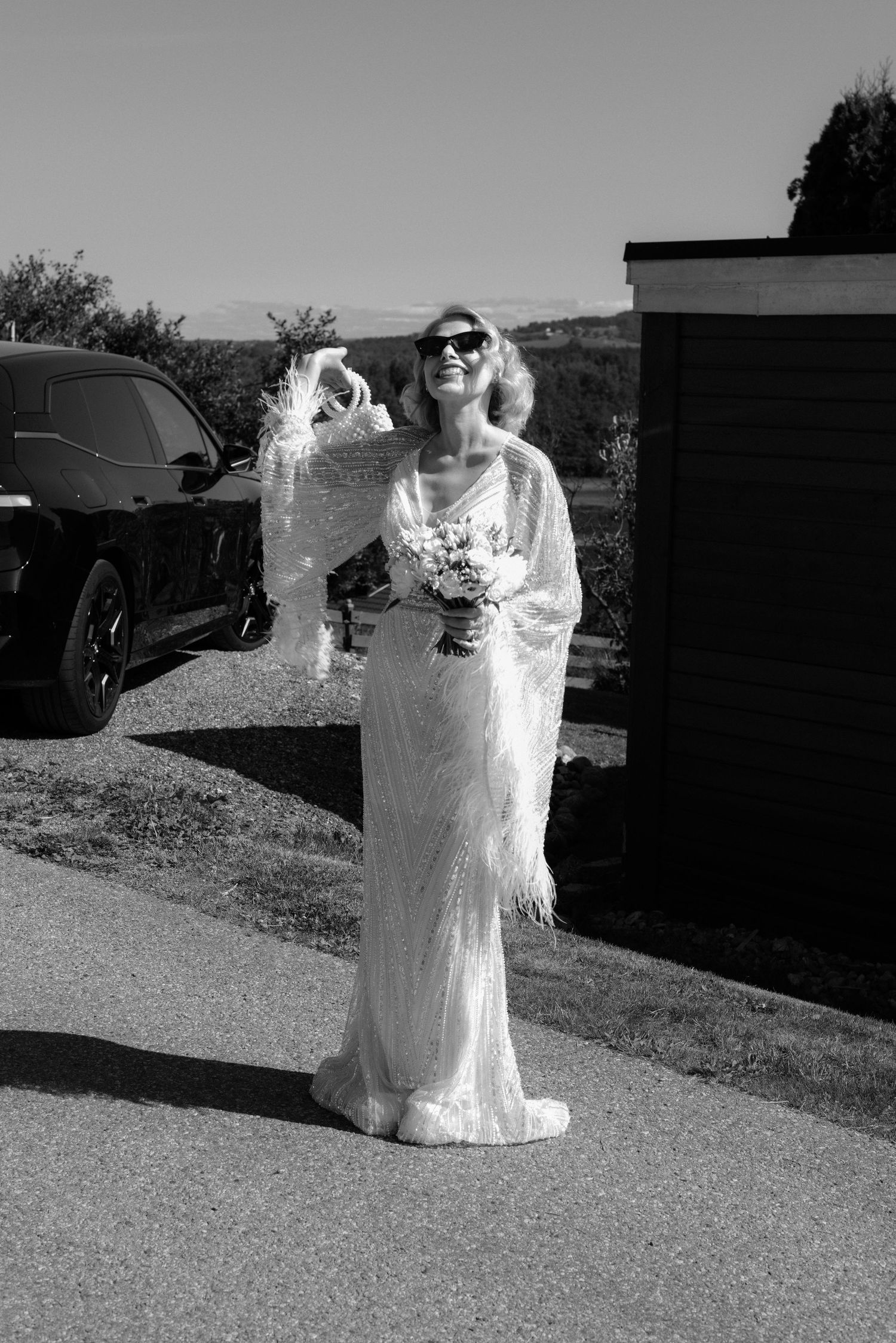 A bride in a vintage-style lace wedding gown and sunglasses poses outdoors in black and white photography.