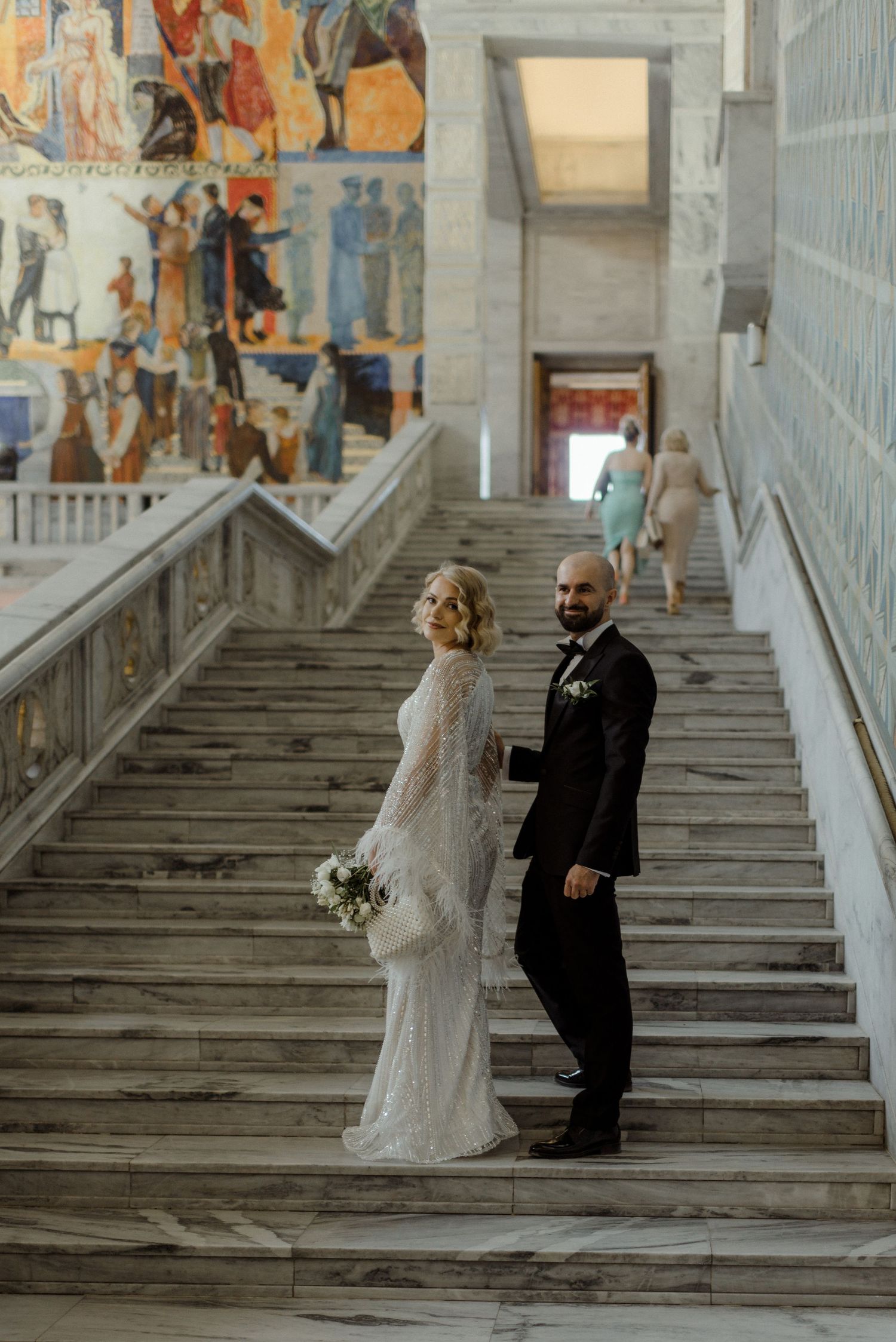 A wedding couple stands on a grand marble staircase with decorative murals adorning the walls in an elegant venue.
