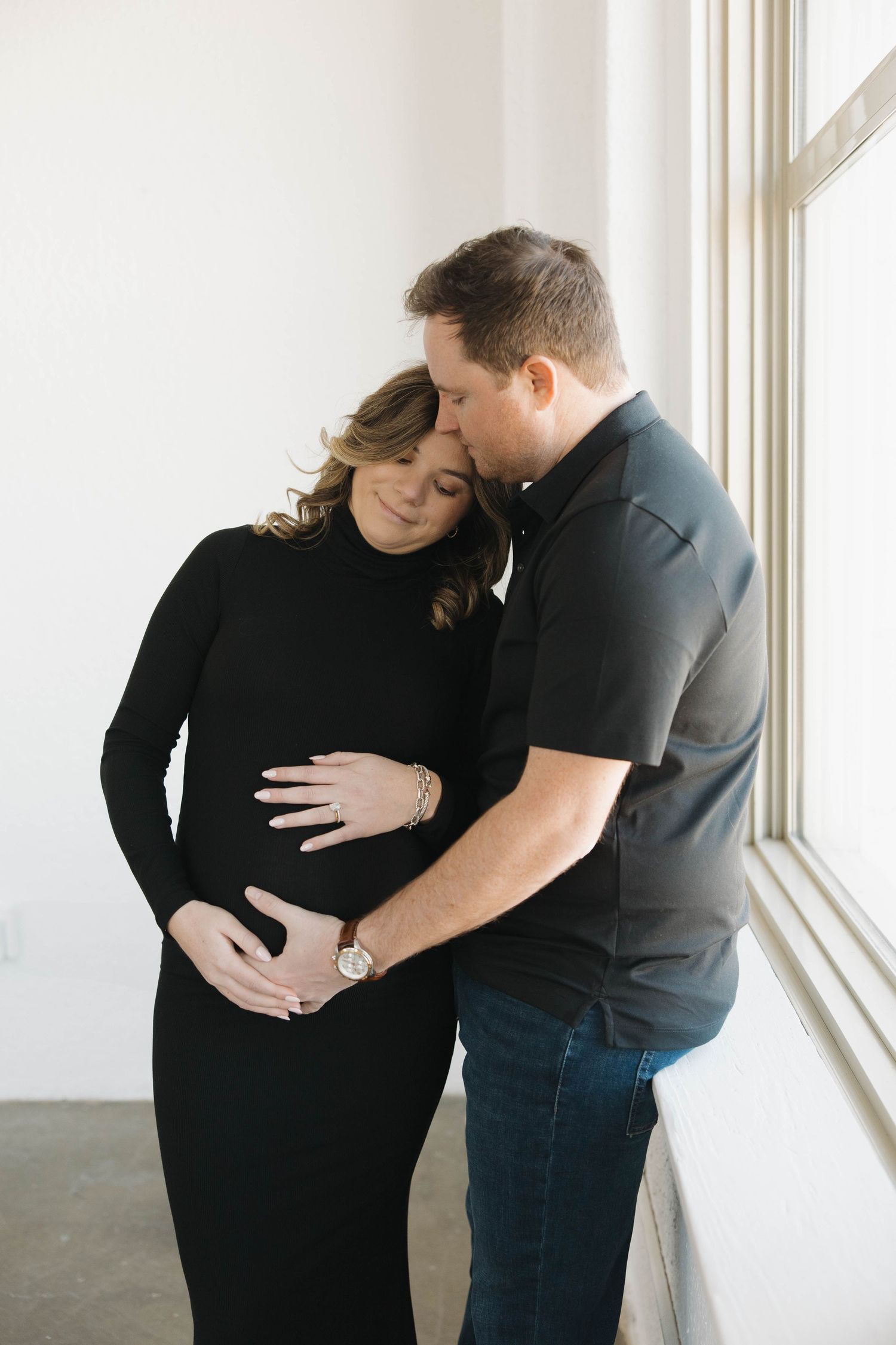 Tender pregnancy photoshoot featuring couple in black outfits near window sharing intimate moment.