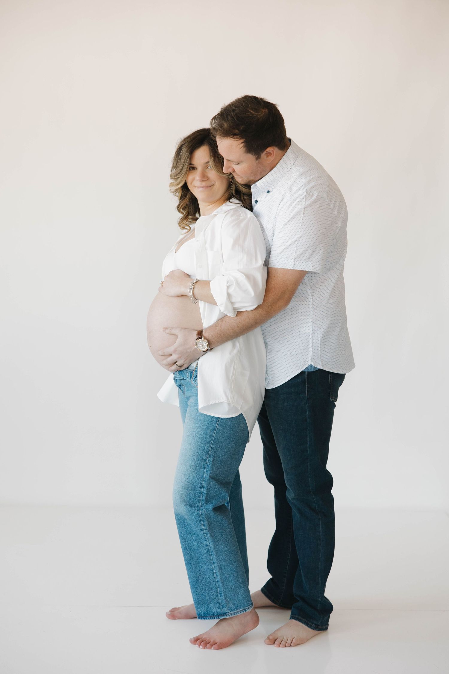Intimate maternity photo of a couple wearing white shirts and jeans embracing against a neutral background.