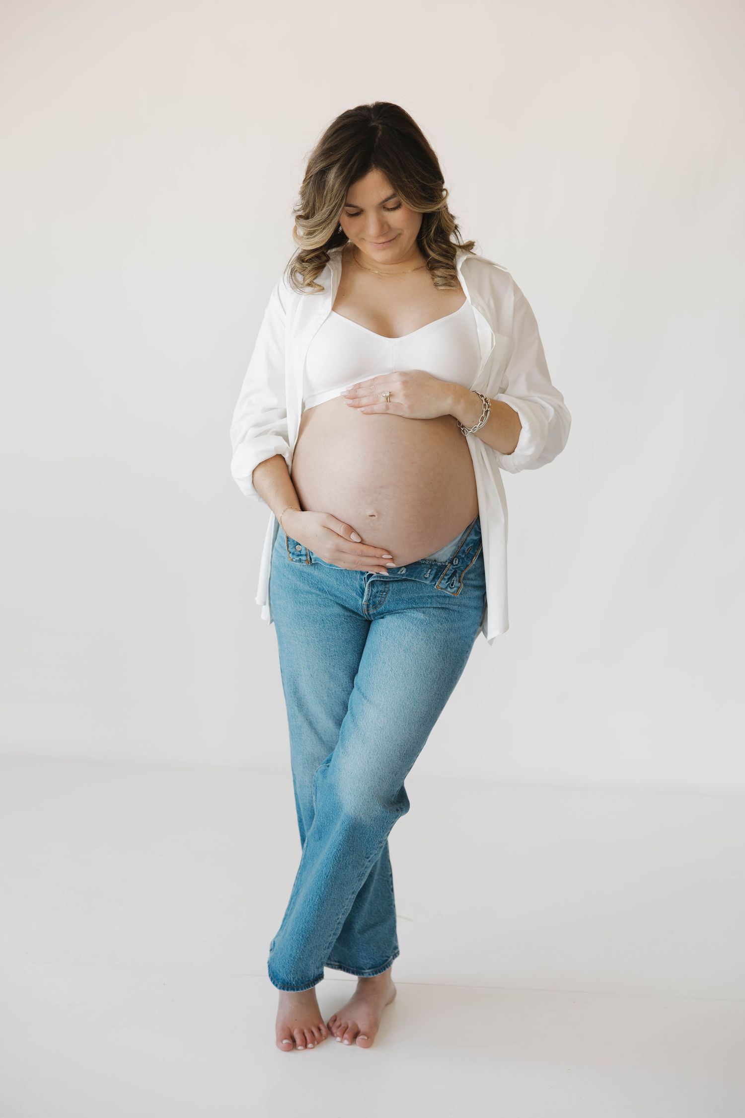 A pregnant woman in jeans and white shirt poses barefoot against a white backdrop.