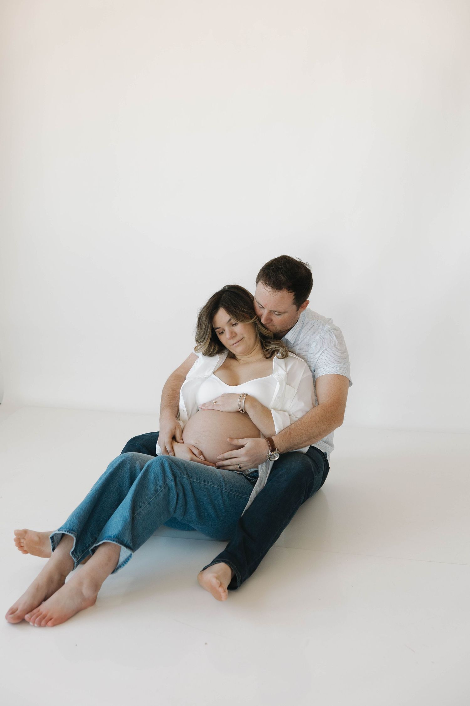 Intimate floor poses of an expecting couple in casual attire against a white background.