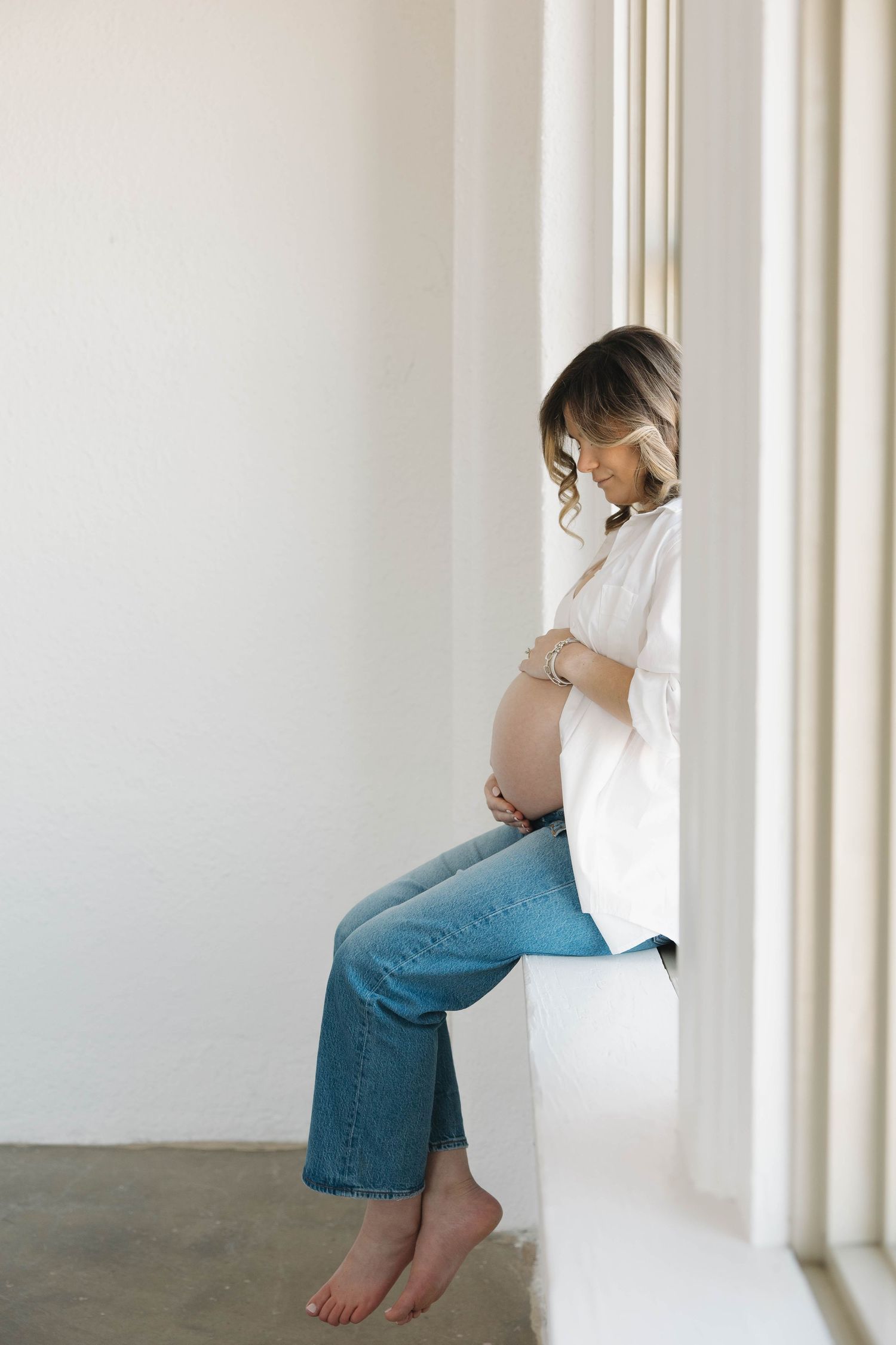 A pregnant woman in jeans and white shirt leans against a white wall near a window.
