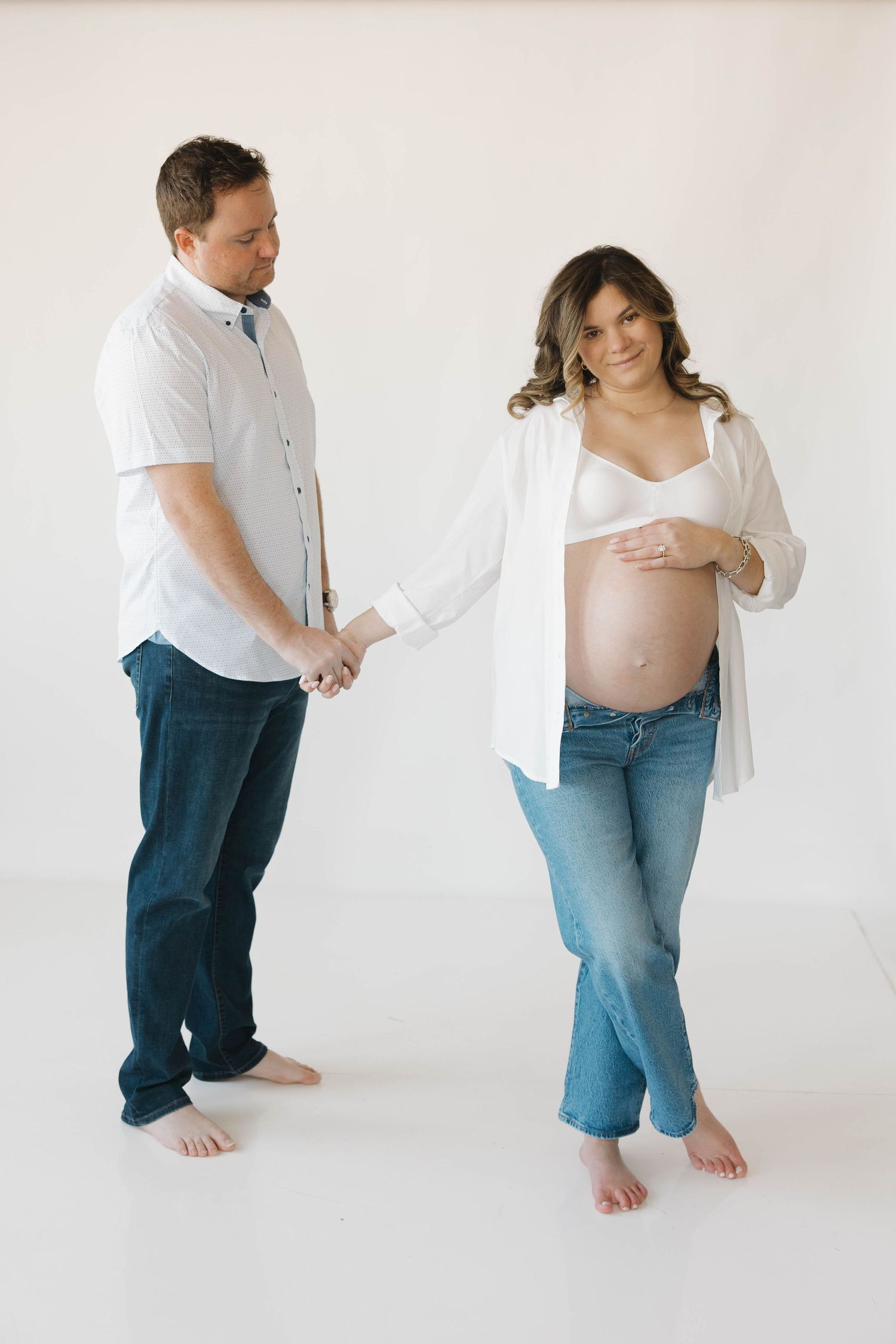 A couple poses together for a maternity photo in casual white clothing against a light backdrop.