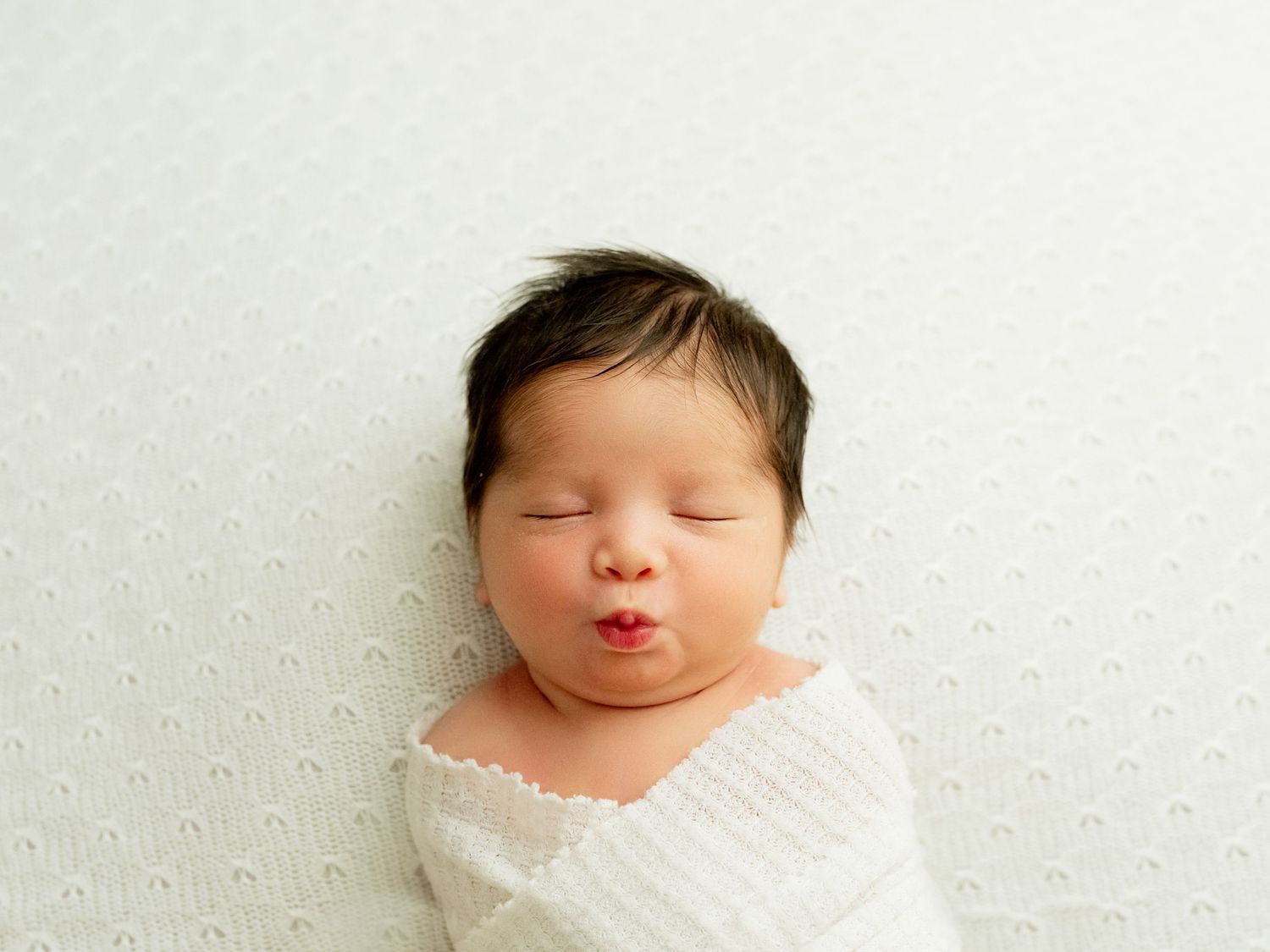 A sleeping infant swaddled in white fabric lying on a white textured background.