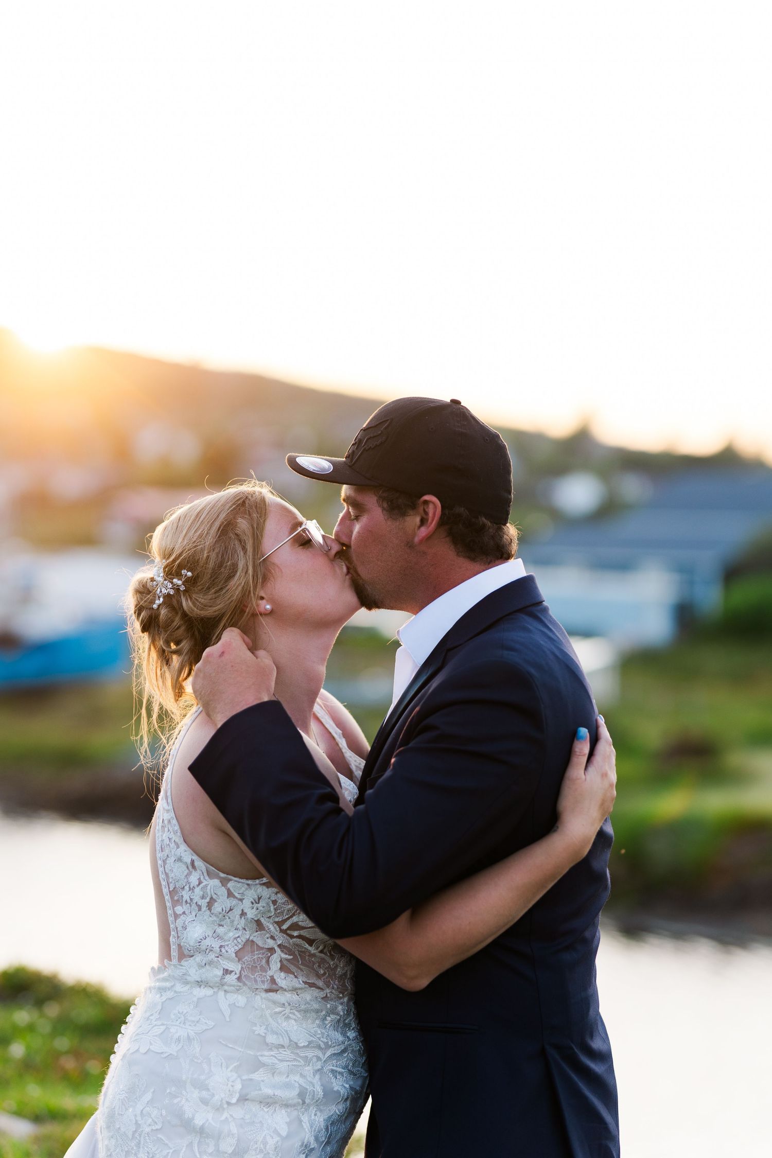 A couple in wedding attire share a romantic kiss at sunset with water and greenery in the background.