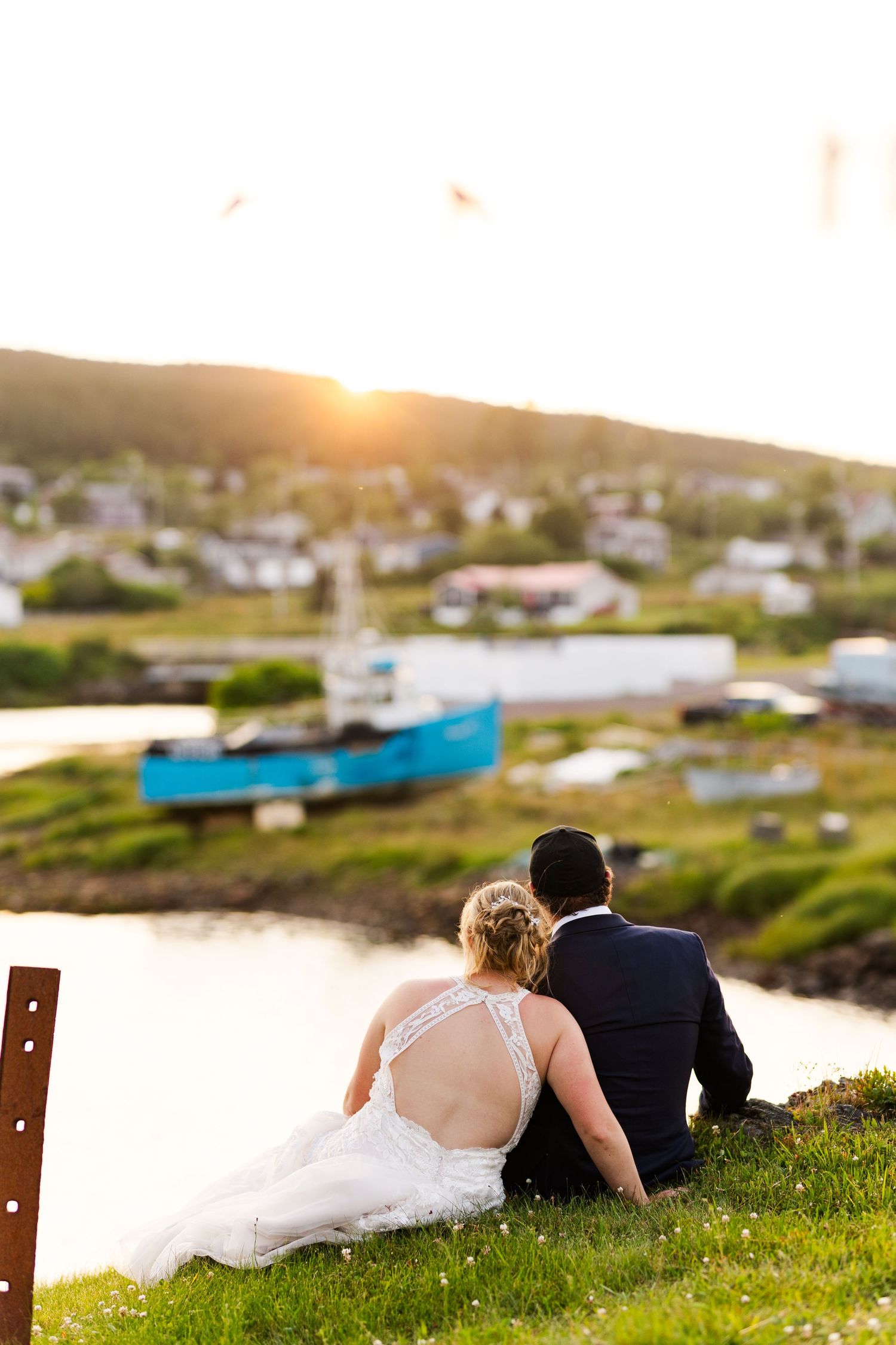 A couple in wedding attire sits on a grassy hill overlooking a coastal village at sunset.