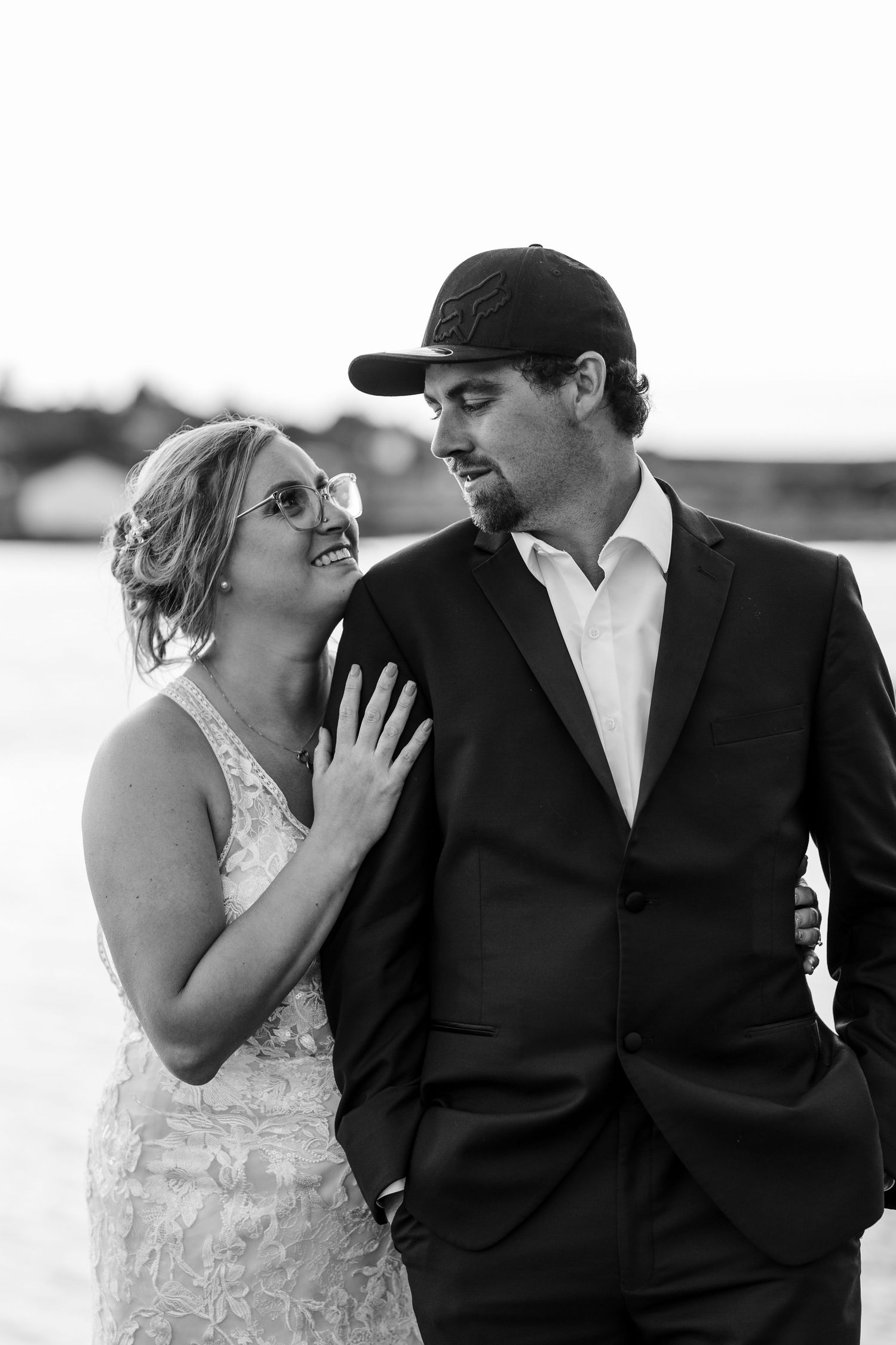 Black and white wedding portrait of a couple sharing an intimate moment outdoors with bride in lace and groom in baseball cap.