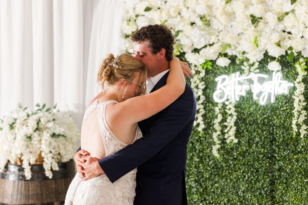 A romantic couple embrace amid elegant white floral arrangements and a green hedge wall backdrop at a wedding reception.